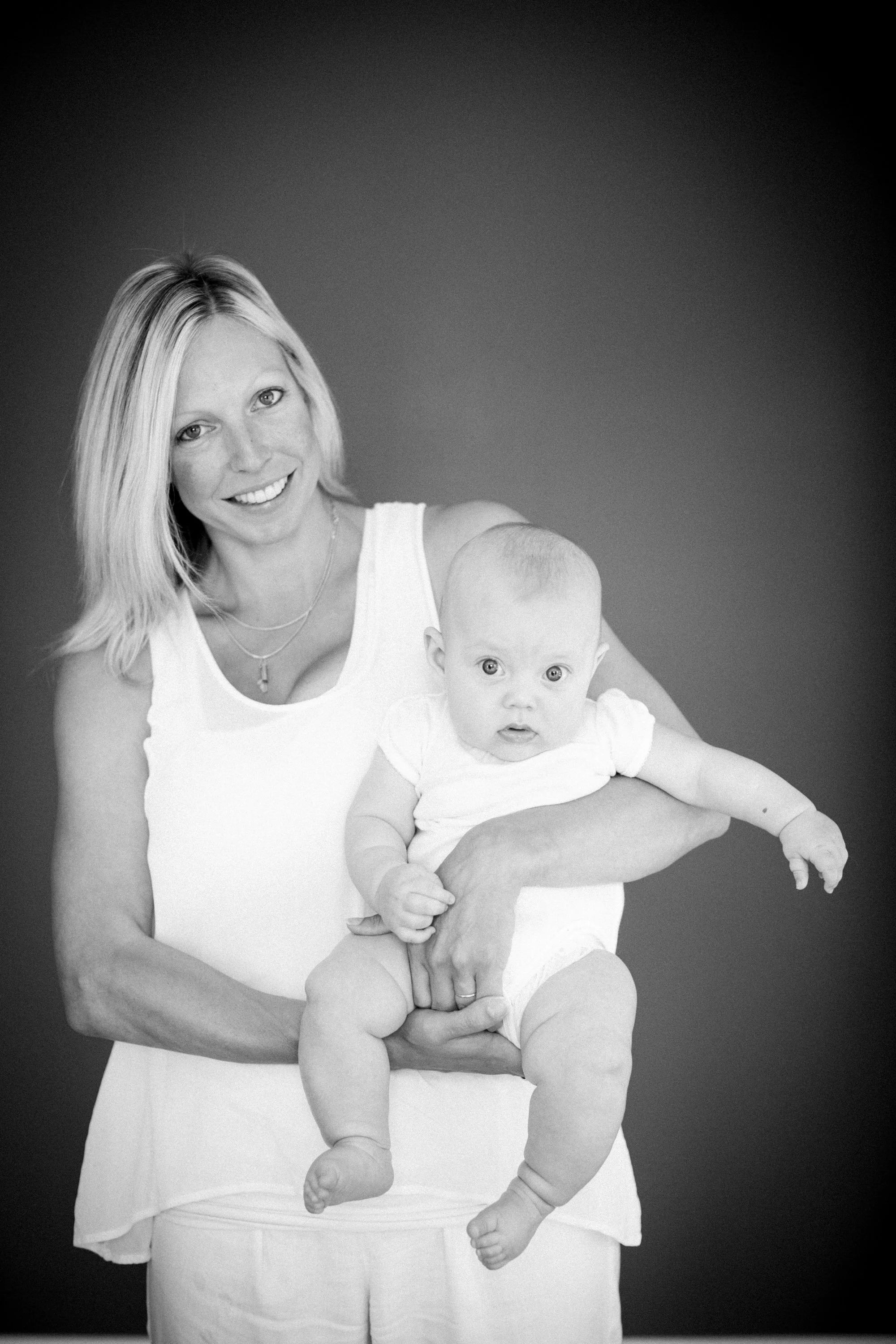 Black and white photo of a smiling woman holding a baby, both facing the camera. The woman has shoulder-length blonde hair, wearing a sleeveless top, and the baby has a surprised expression, wearing a light-colored outfit.