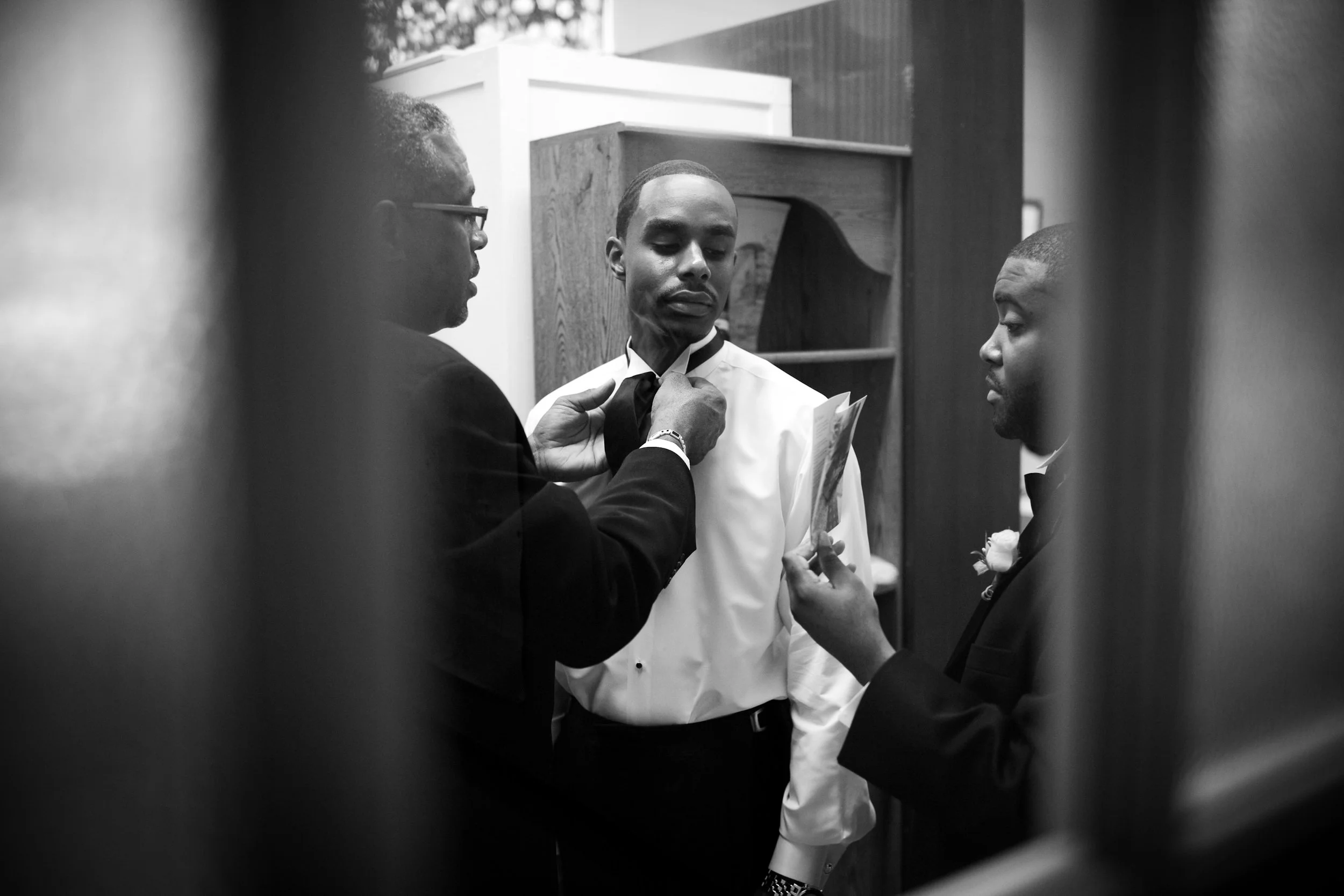 A black and white photo of three men in formal attire, one adjusting the collar of a man in a white shirt, with the third man holding a card, seen through a narrow gap.