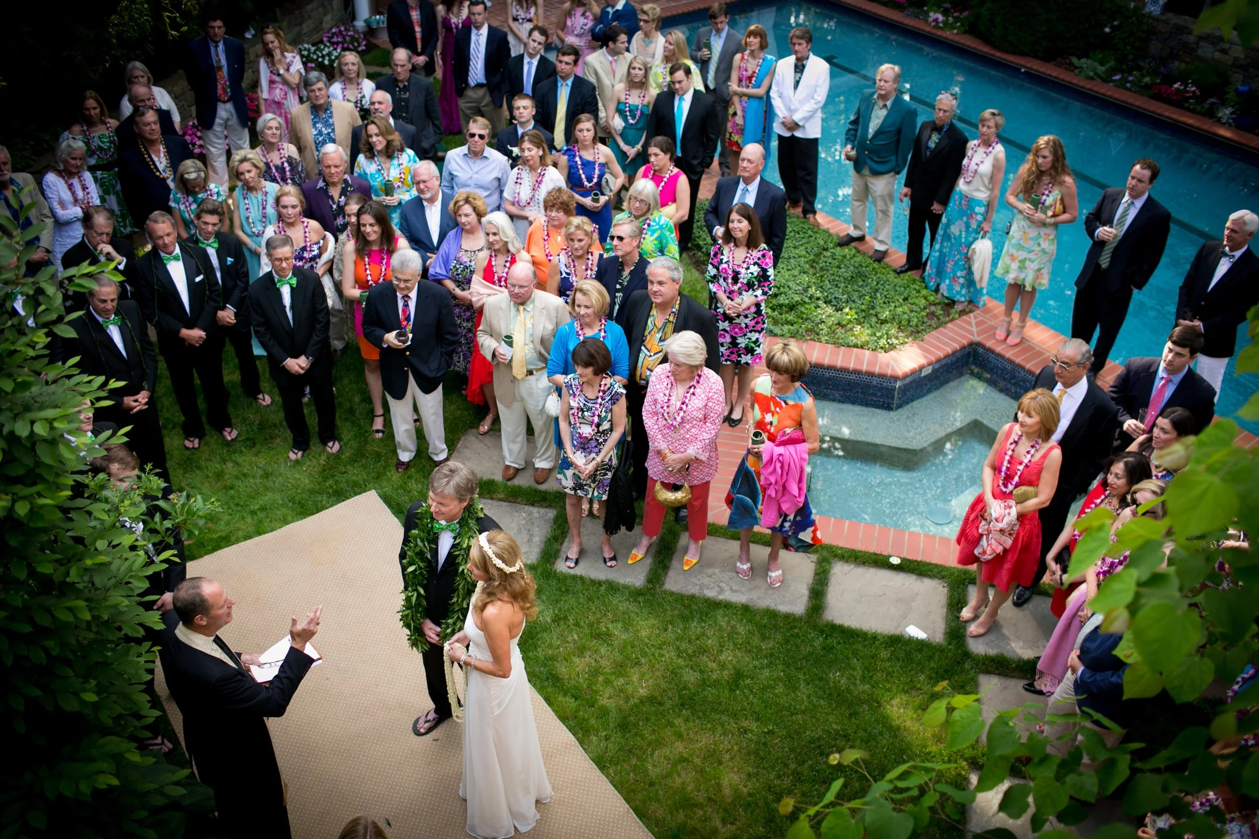 A wedding ceremony taking place outdoors with a bride and groom standing on a small platform in front of a man officiating. The bride wears a white dress and floral headband, and the groom wears a suit with a lei. Guests gather around, dressed in col