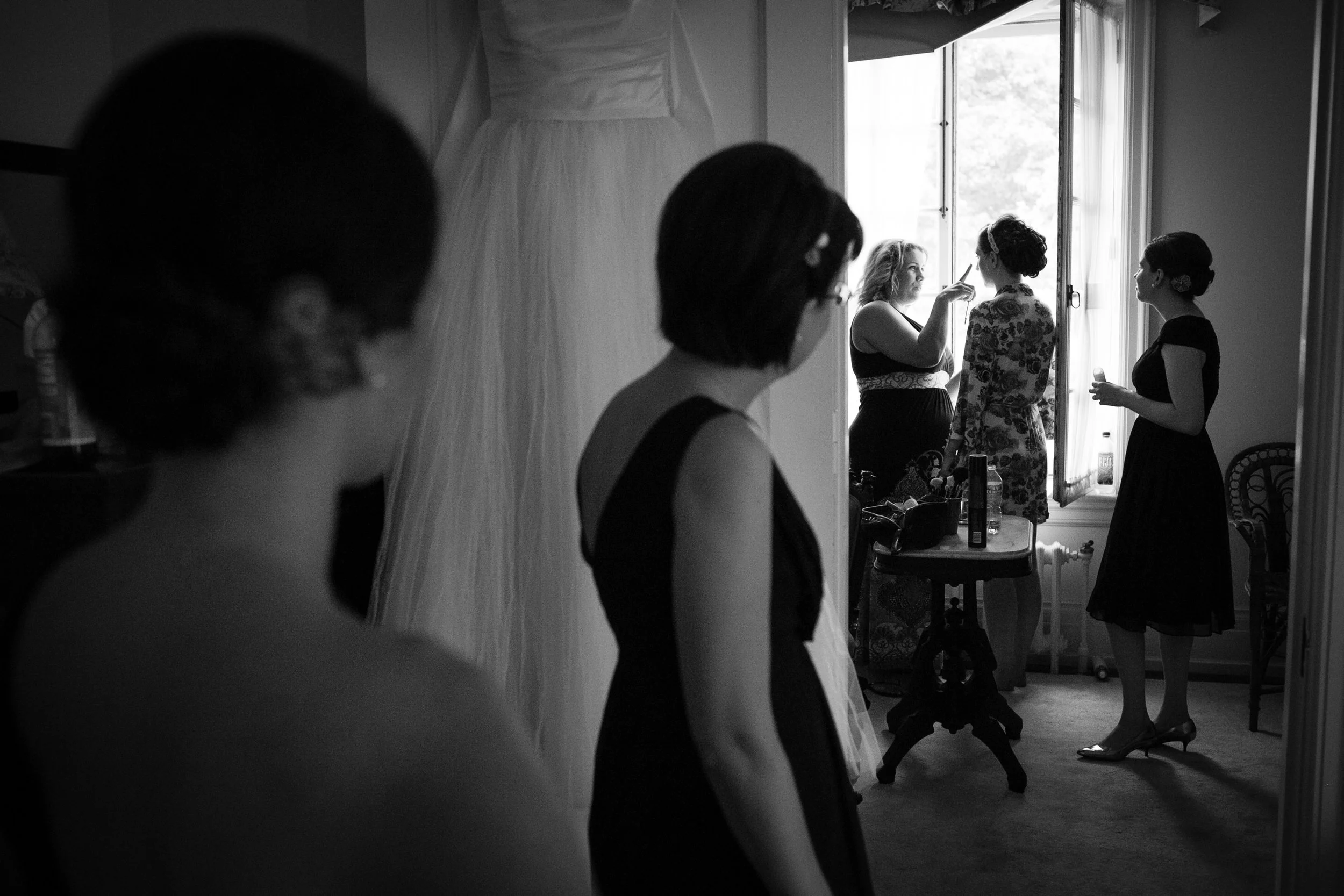 Four women in a room preparing for a special event, possibly a wedding, with one woman applying makeup to another woman near a window.