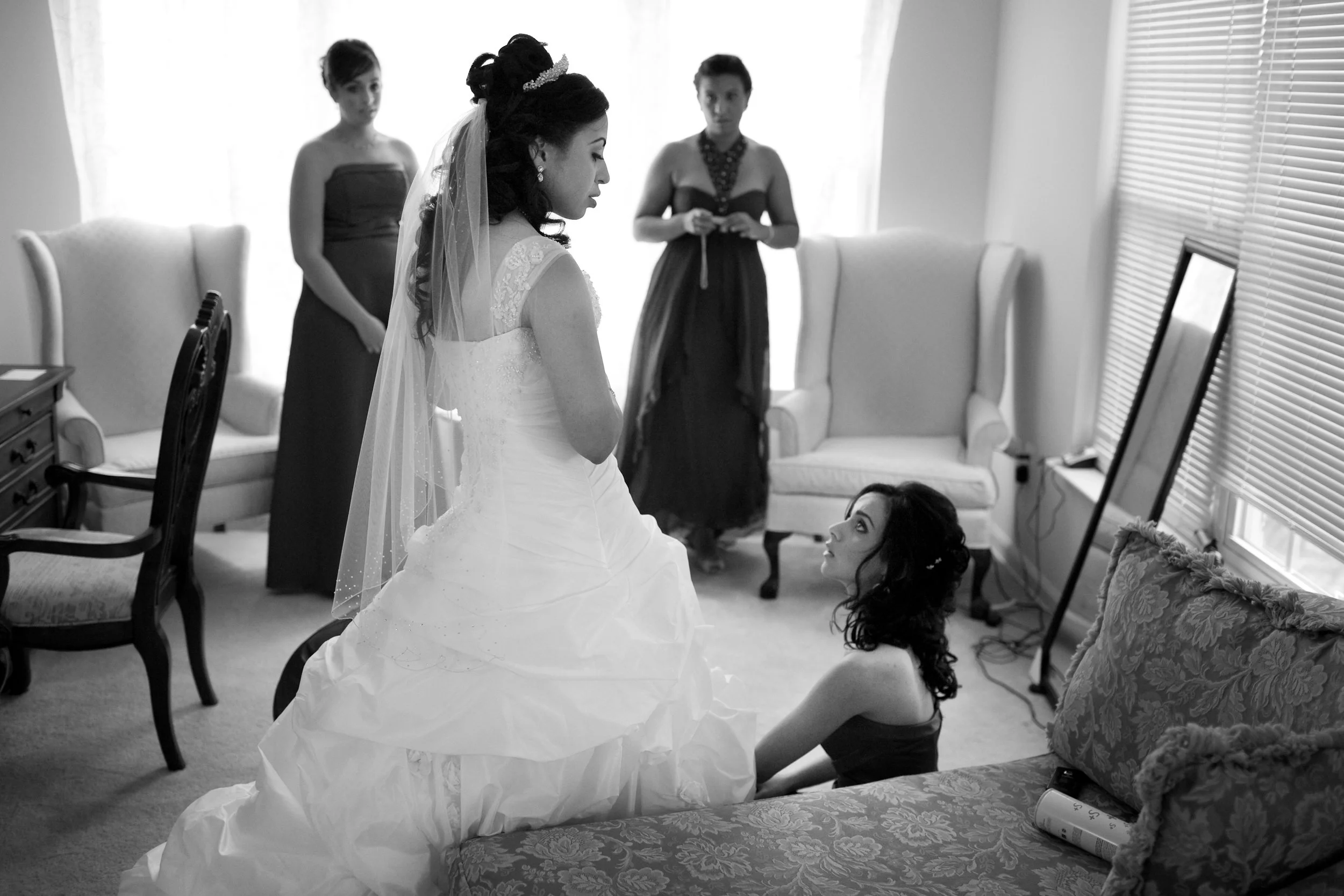 A bride in a wedding dress and veil looks down at a seated woman in a dark dress, with two other women in the background in a room with large windows and blinds.