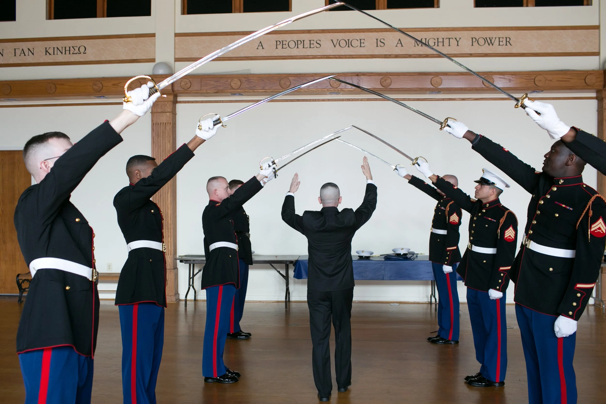 Military band performing a sword salute with drummers in uniform and a conductor in black suit in a hall.