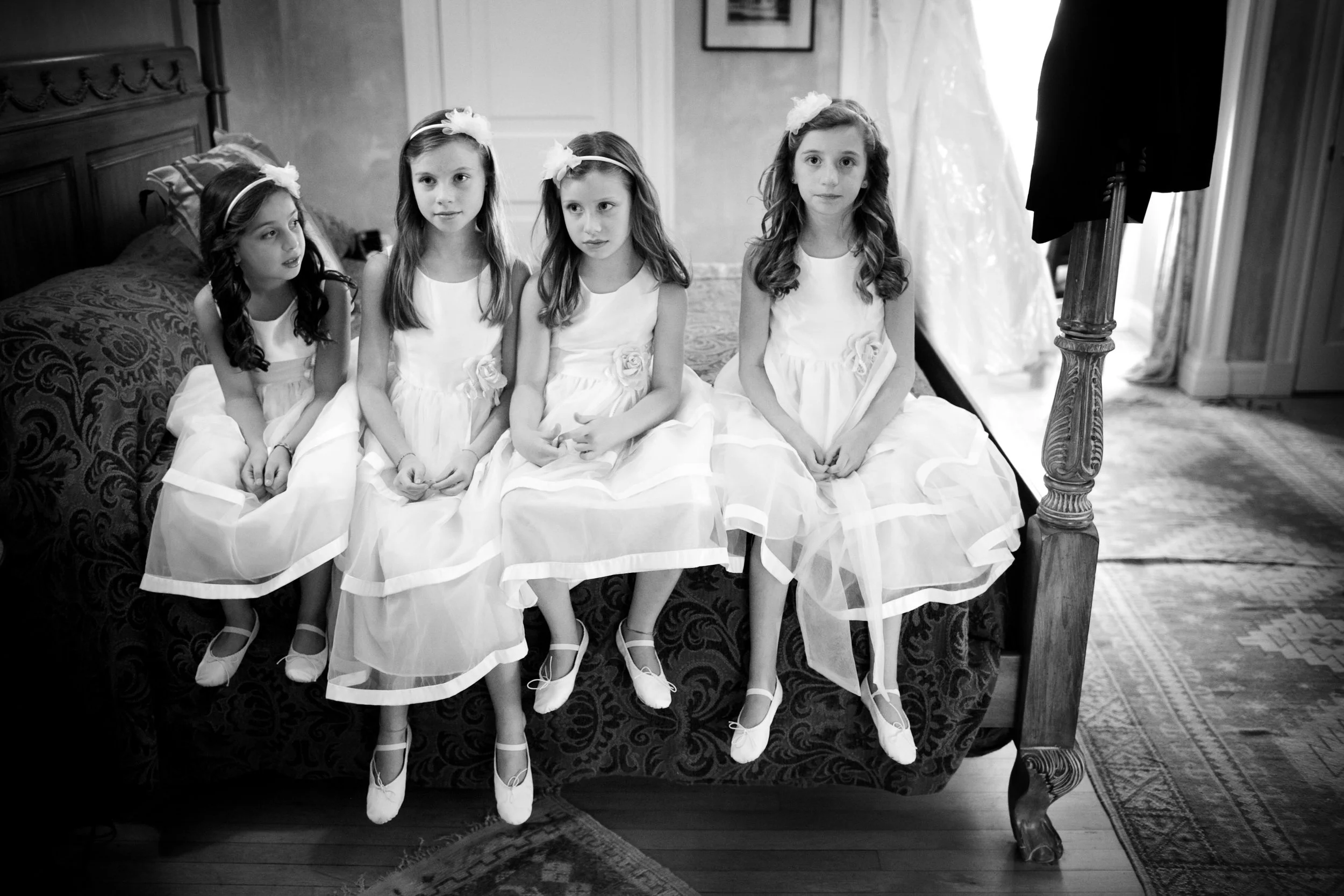 Four young girls in white dresses and ballet slippers sitting on a bed, preparing for a formal event, with a wedding dress visible in the background.