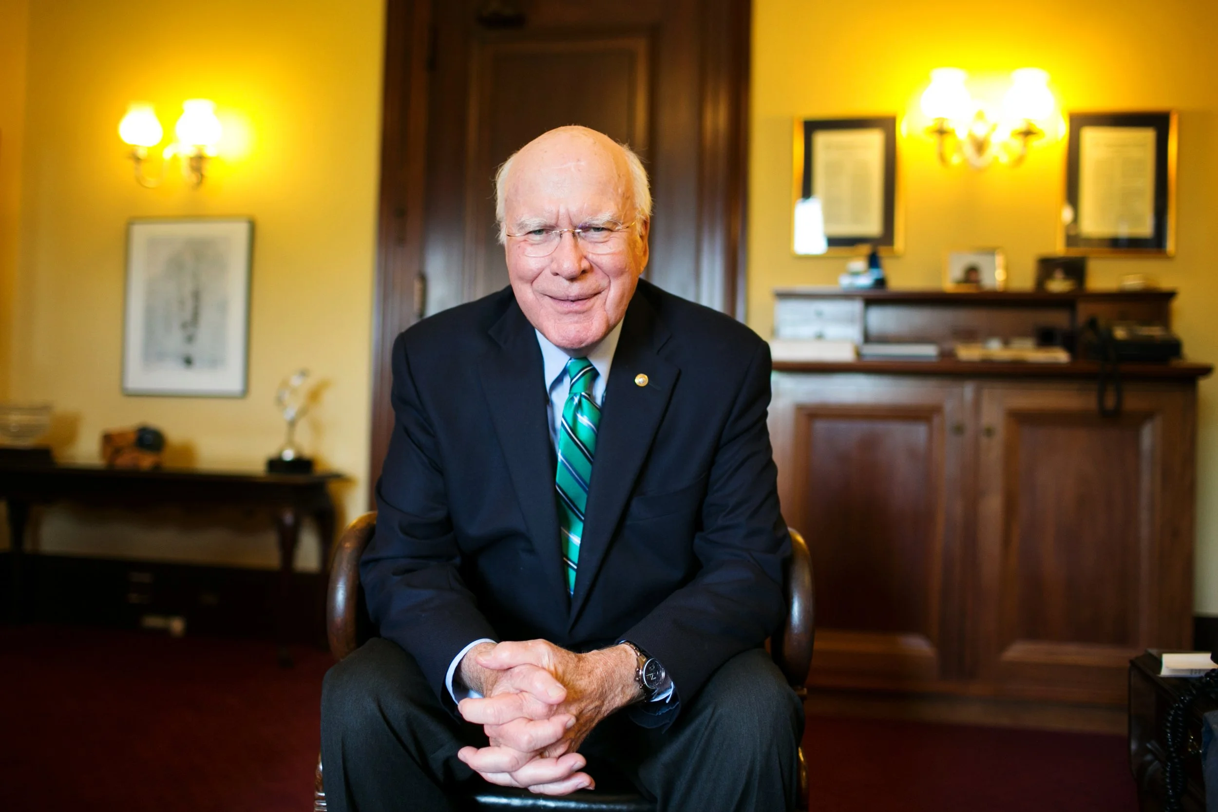 An elderly man in a dark suit and striped green tie sitting in an office, smiling at the camera. The background features wooden furniture, yellow walls, and framed pictures with warm lighting.