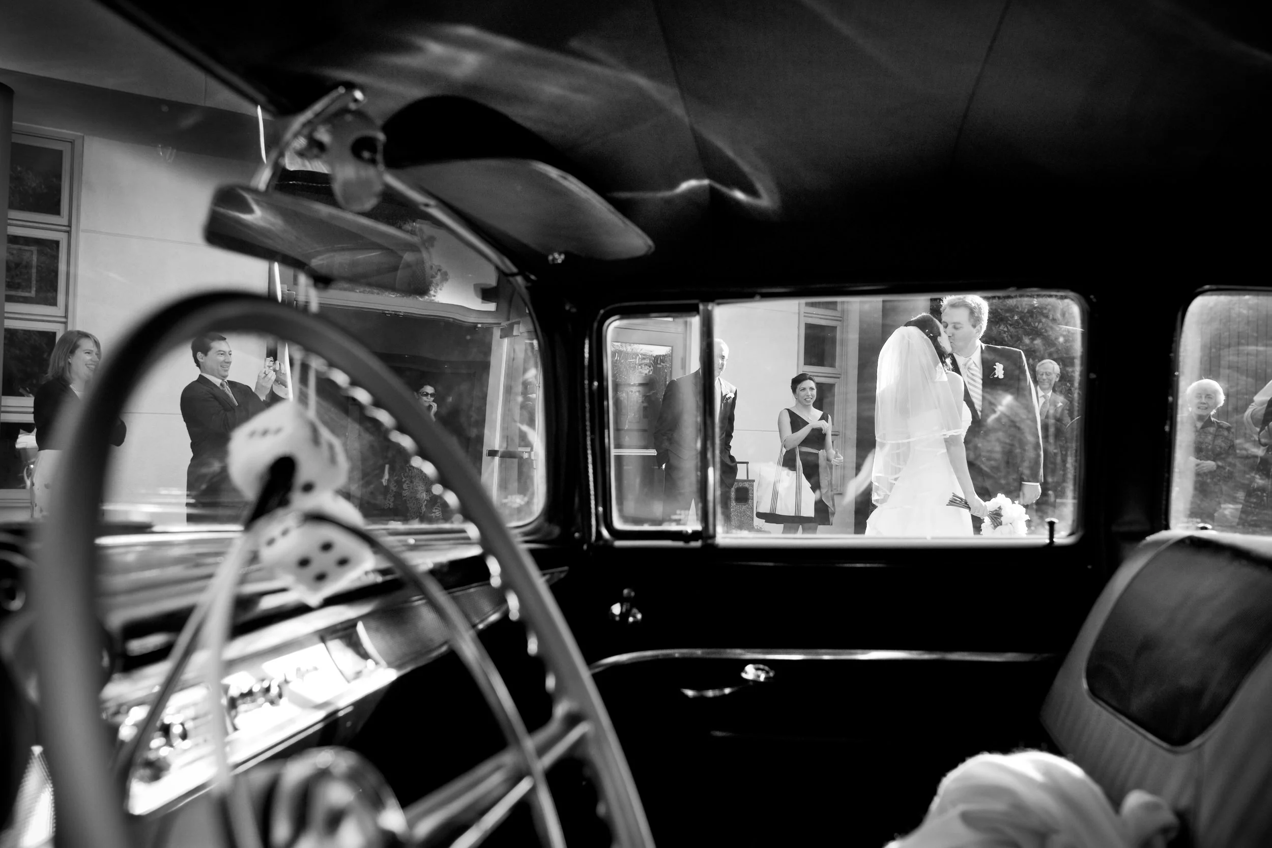 A black and white photo taken from inside a vehicle showing a bride and groom kissing outside, with wedding guests standing nearby.