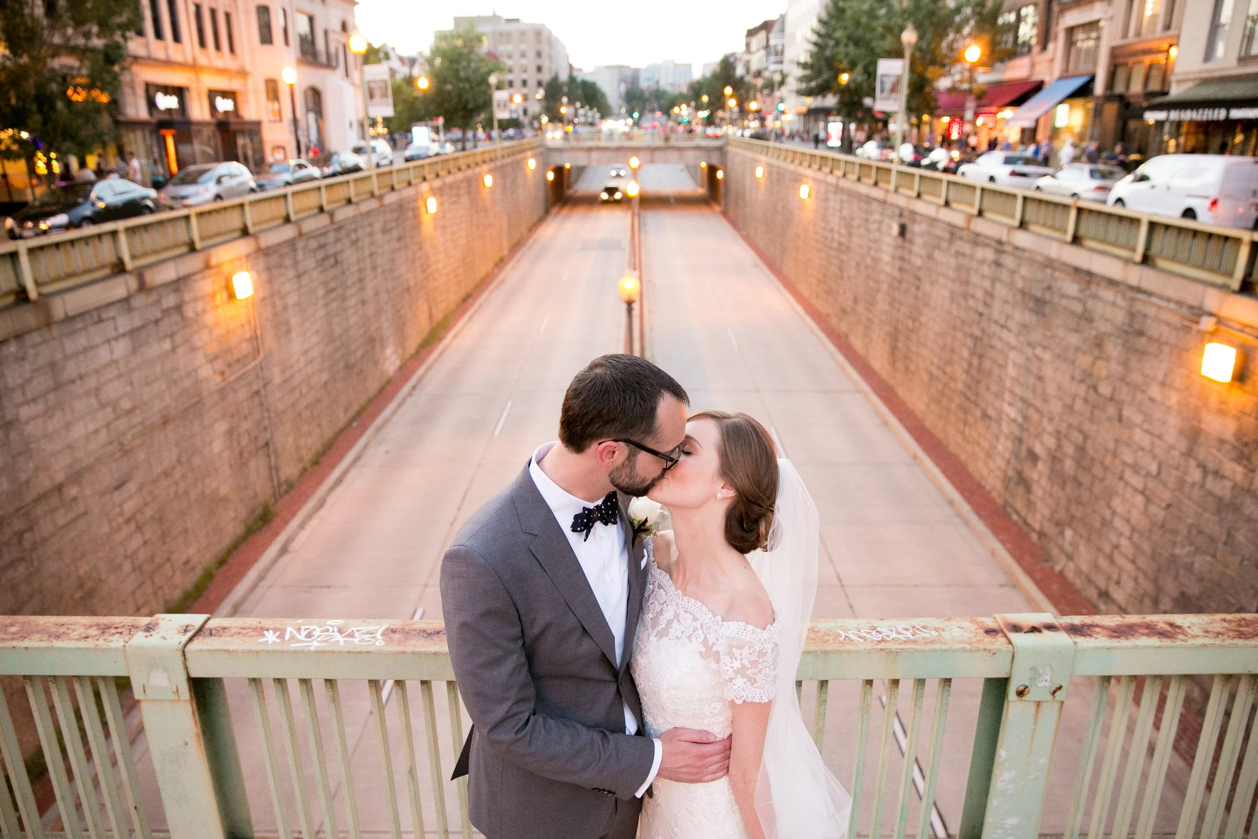 A bride and groom kissing on a bridge over a sunken street at dusk in an urban setting.