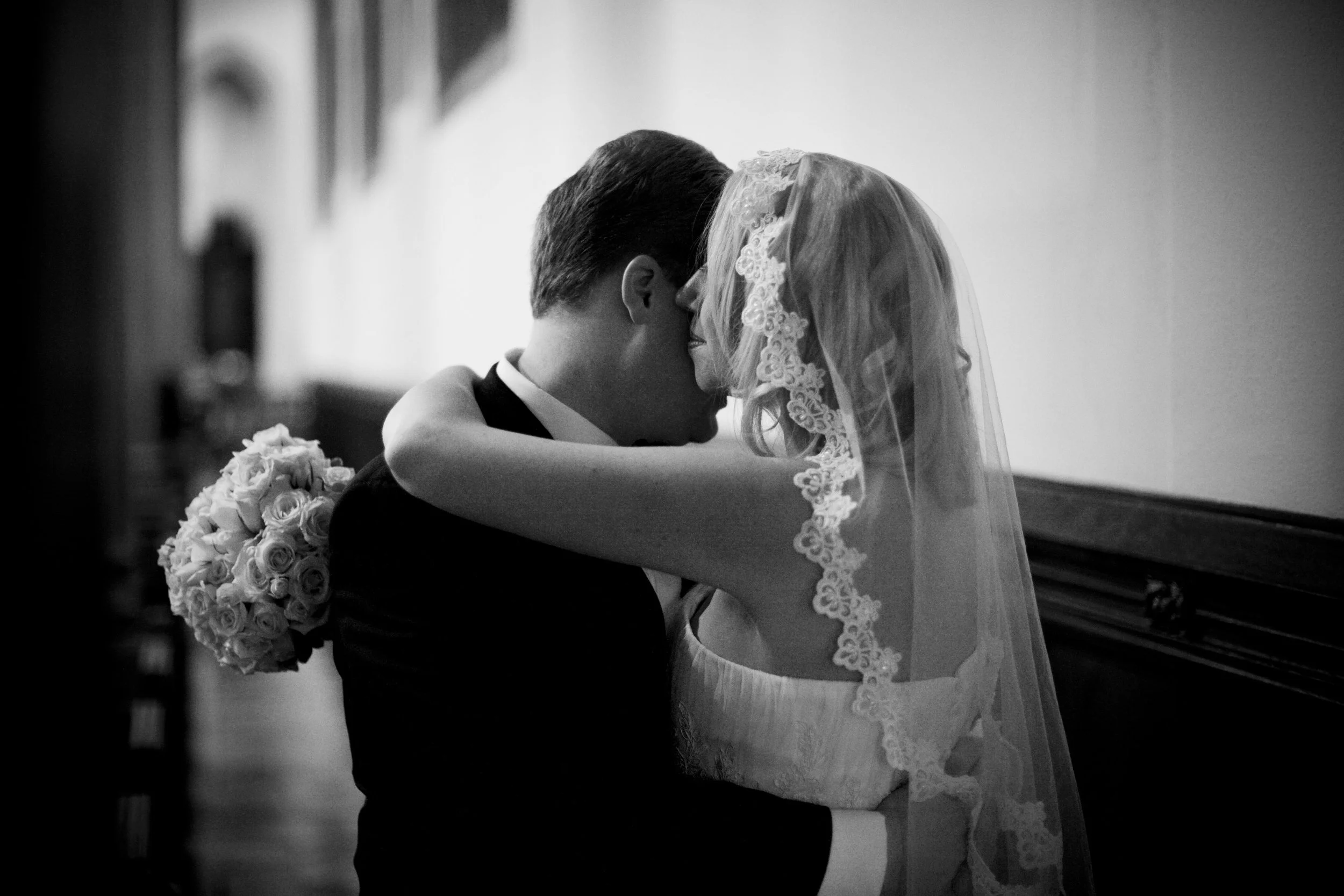 Black and white photo of a bride and groom sharing a kiss. The bride is wearing a lace veil and has her arms around the groom's neck. The groom is in a tuxedo and holding a bouquet of roses. They are indoors, standing close together.