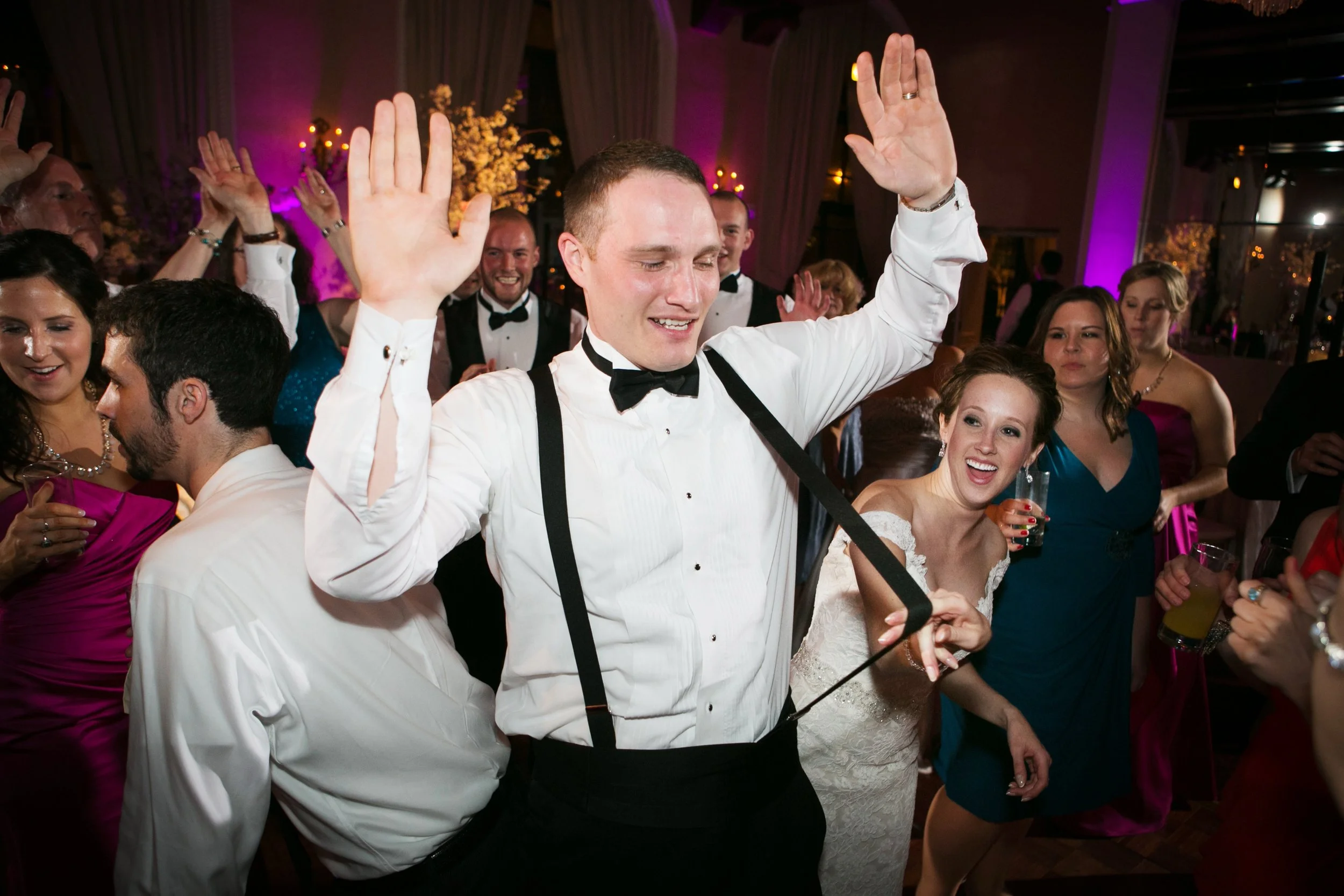 People dancing and celebrating at a wedding reception, with the groom in a white shirt and black suspenders in the foreground raising his hands.
