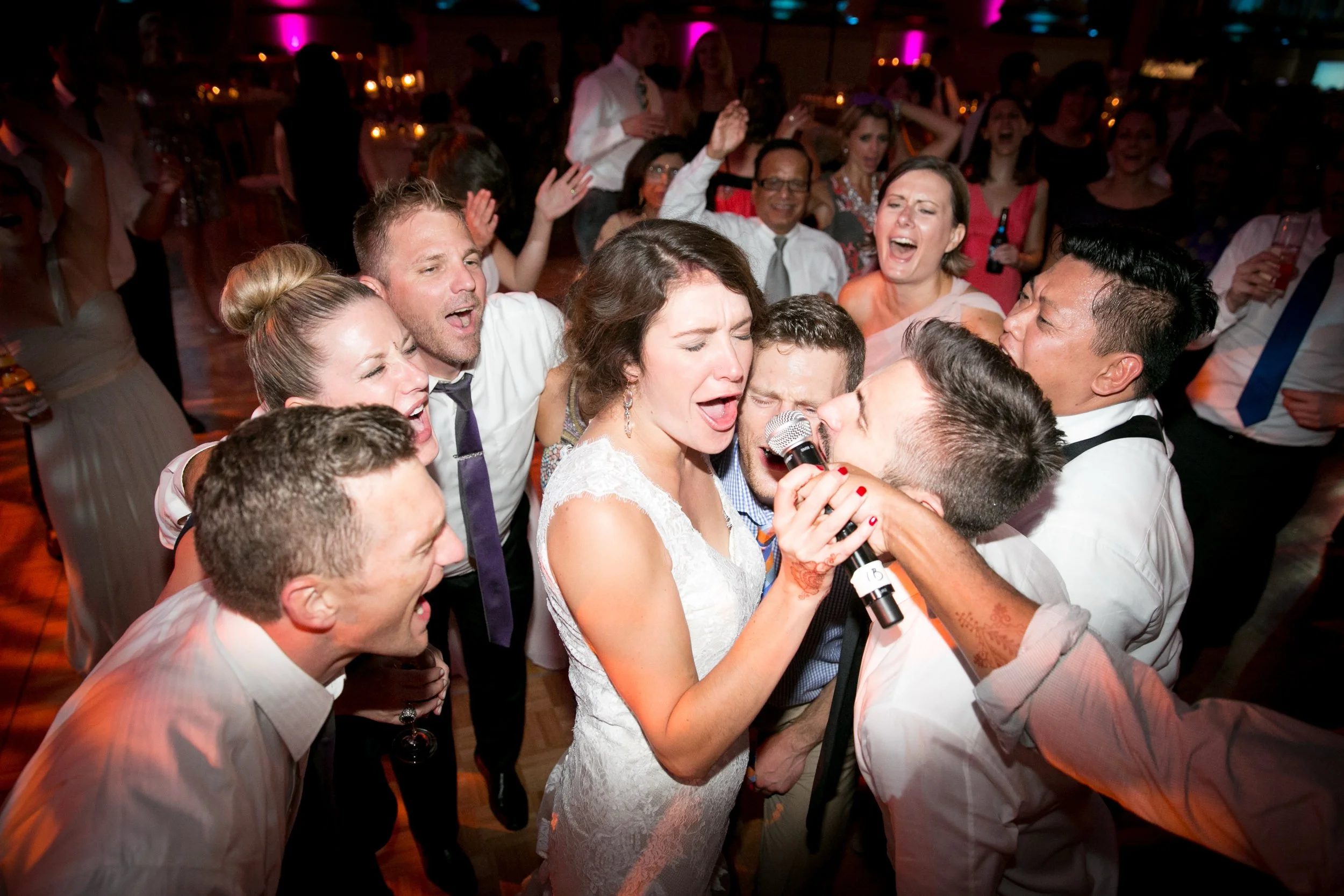 People at a wedding reception singing and dancing together, with a woman in a white dress holding a microphone.