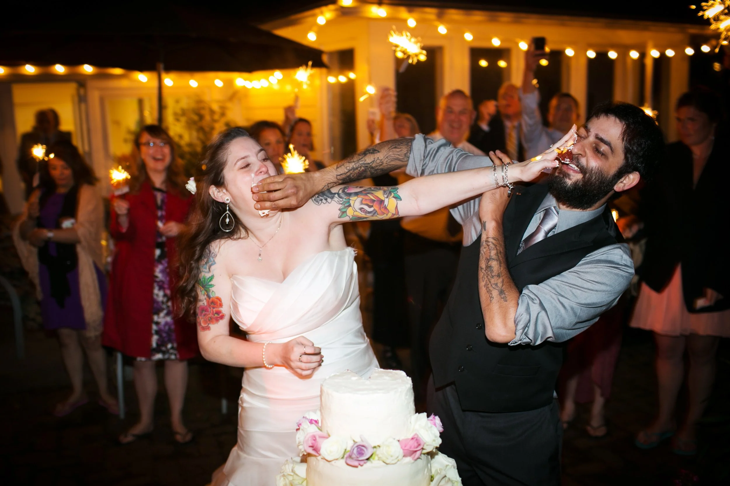 Bride and groom celebrating at their wedding reception with guests in the background, cake on the table, and sparklers in the night sky.