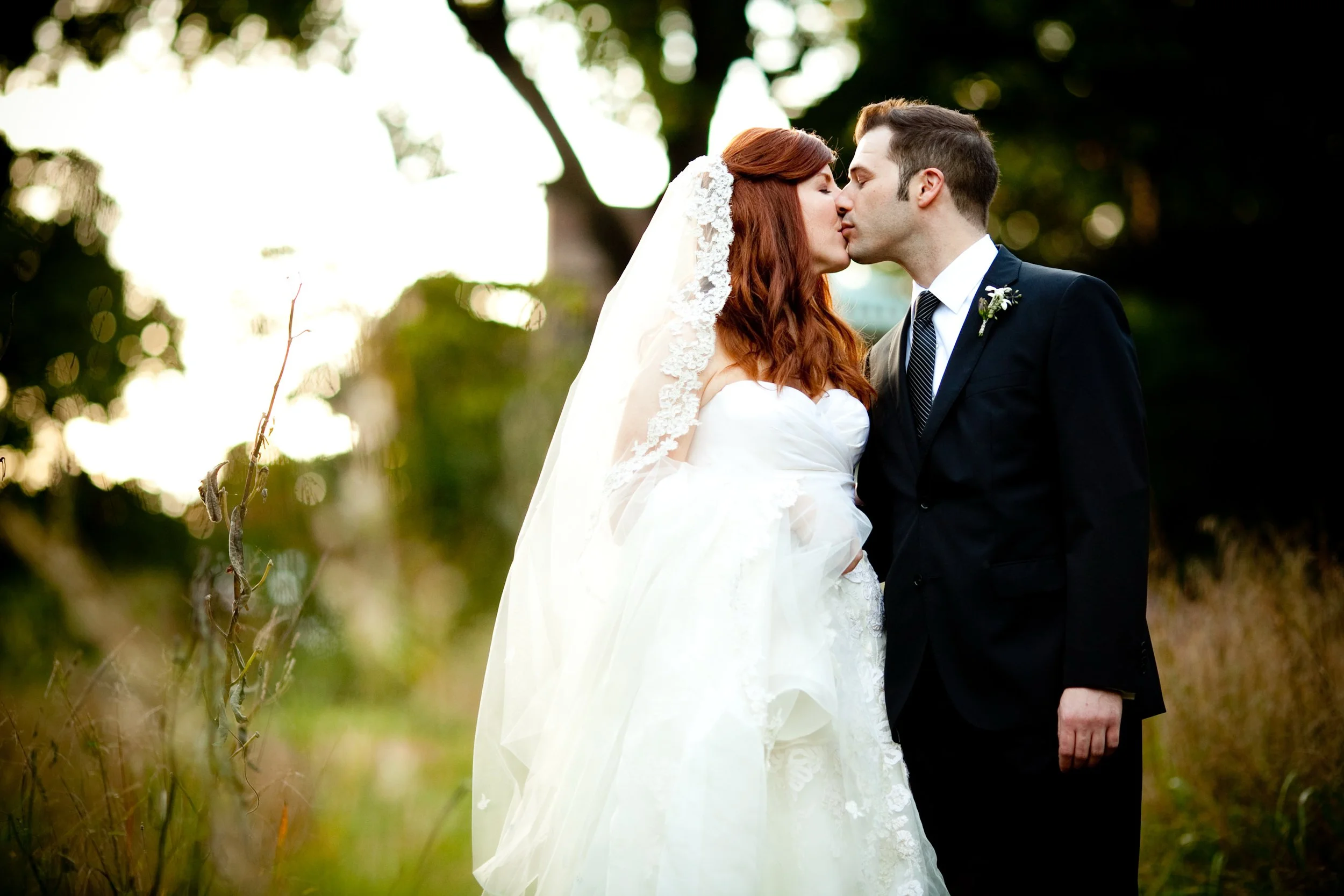 A bride and groom kiss outdoors in a natural setting during their wedding, with trees and greenery in the background.