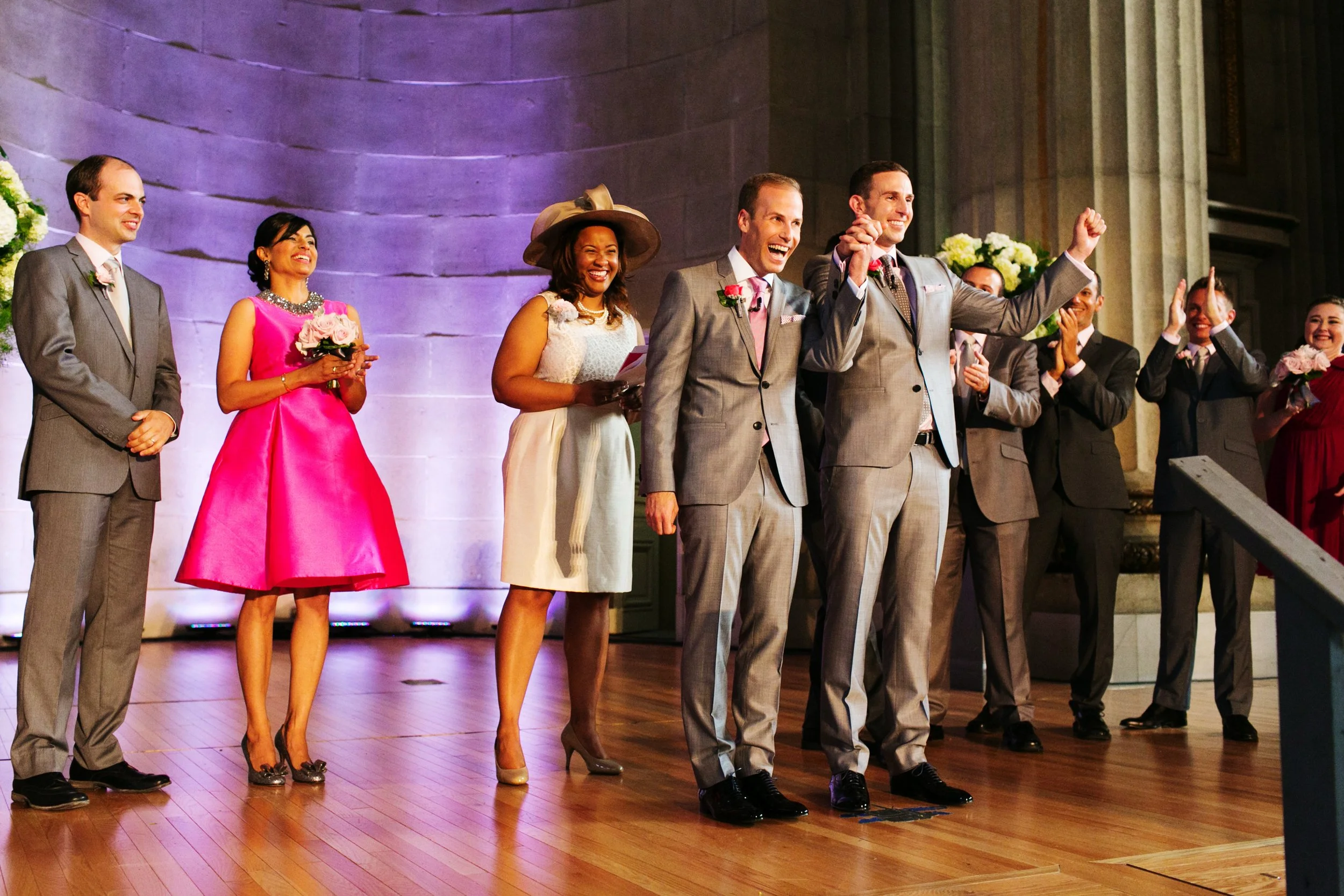 A wedding celebration with a group of people on stage, including groomsmen and bridesmaids, some holding bouquets, with a couple in the center joyfully celebrating.