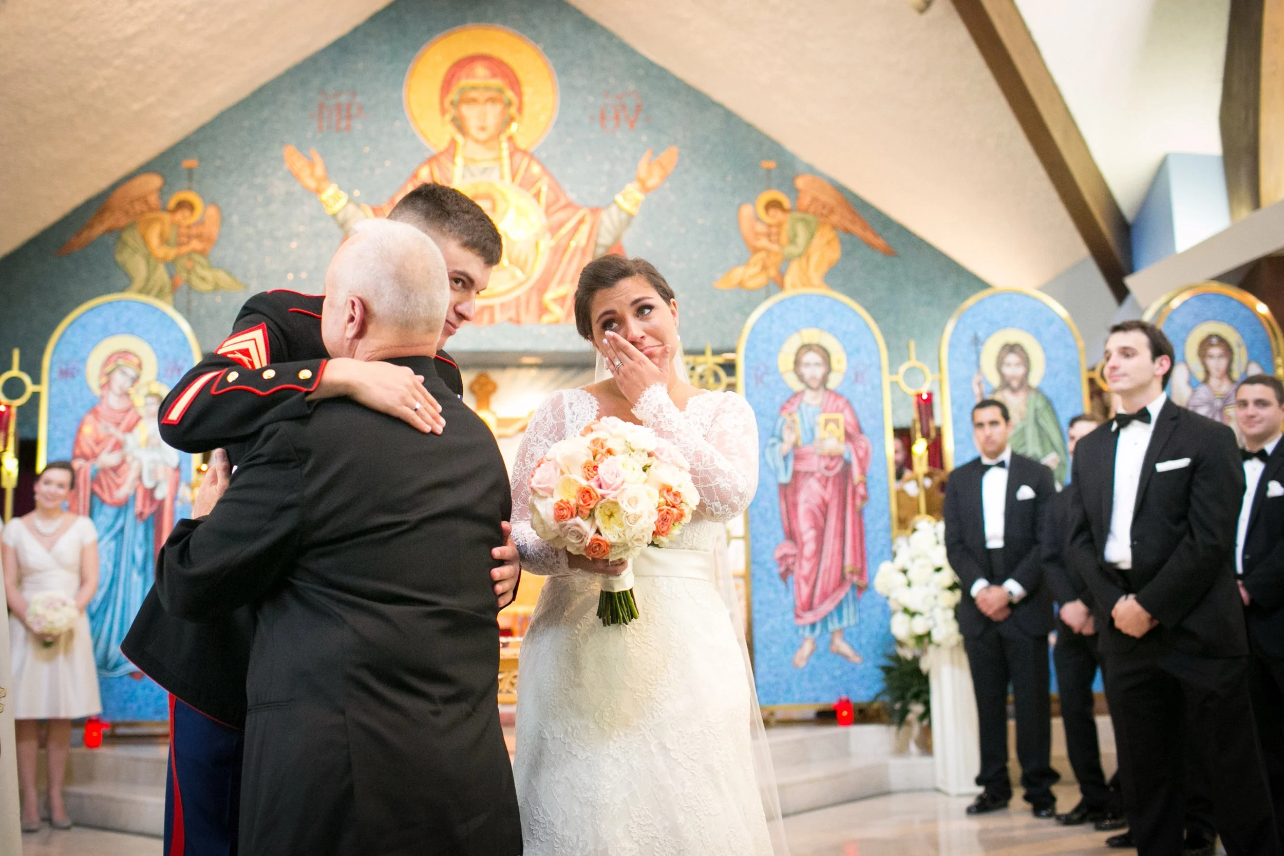 A bride with a bouquet of white, pink, and peach roses is emotional during her wedding ceremony in a church. She is hugging an older man in a military uniform while a young man in a military uniform looks on, and other men in tuxedos stand nearby. Th