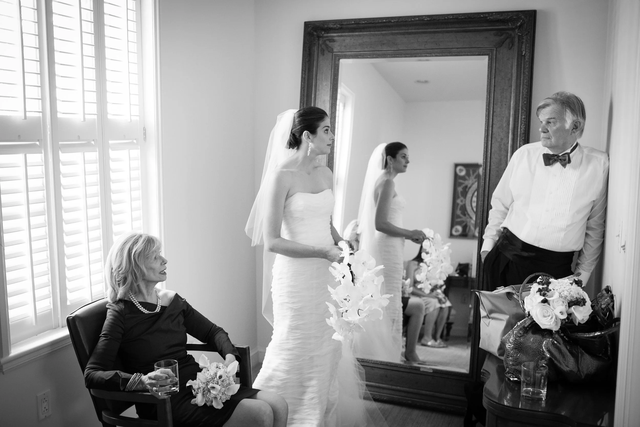A bride in a wedding dress and veil holding flowers standing in front of a large mirror. An older woman in black attire holding a glass and a bouquet sitting nearby. A man in a white shirt with a bow tie leaning against the wall, with a bouquet on a 