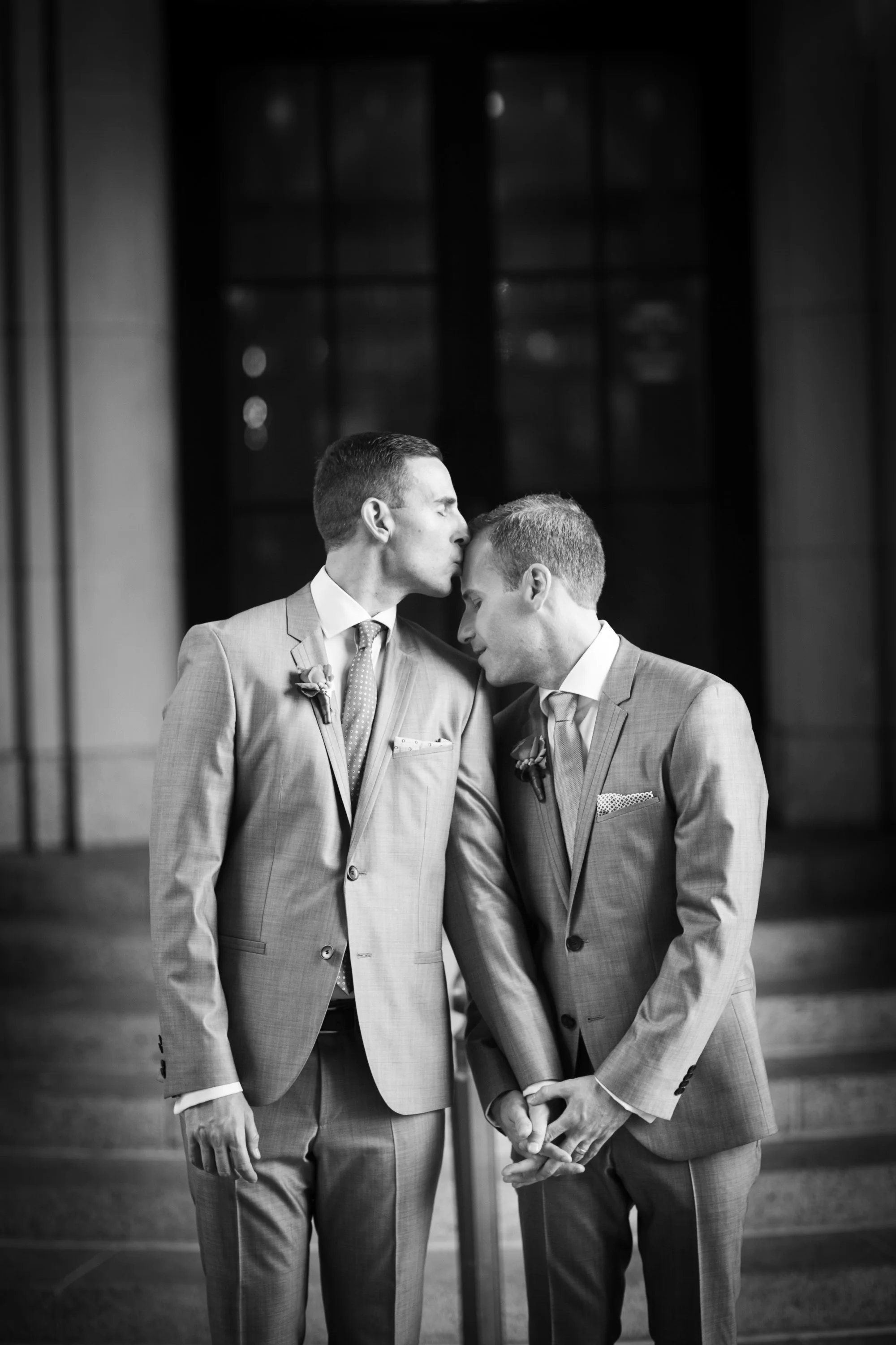 Two men in matching suits are holding hands in front of a large window, with one leaning in to kiss the other's forehead, in a black and white photograph.