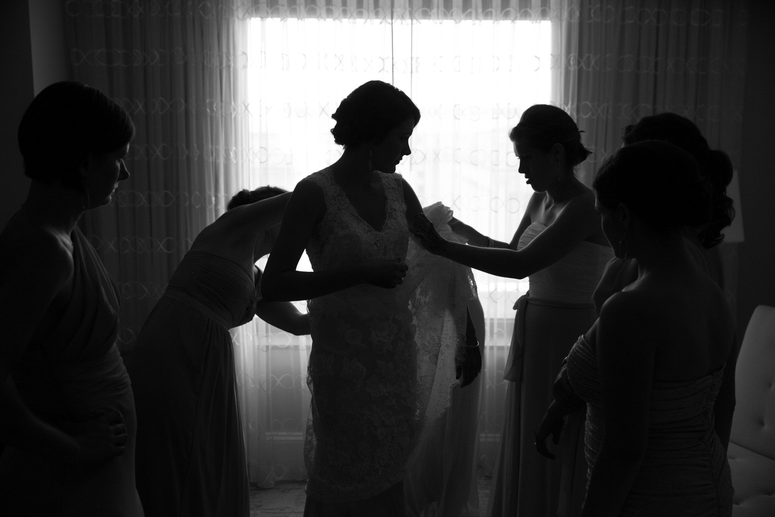 Silhouette of a bride surrounded by her bridesmaids as they help her prepare for her wedding in front of a backlit window.