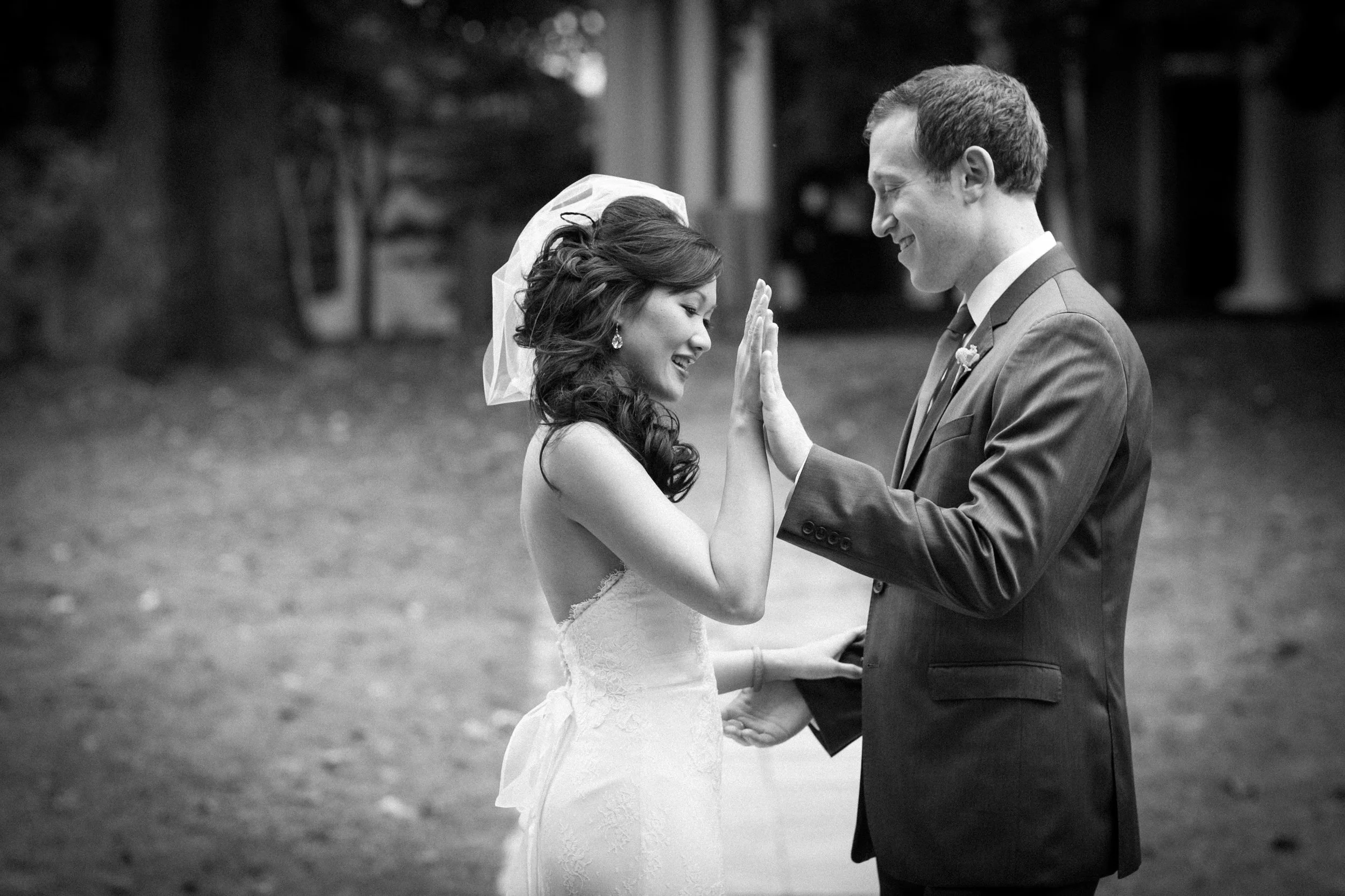 A black and white photo of a bride and groom facing each other, smiling, and touching hands during a wedding ceremony outdoors.