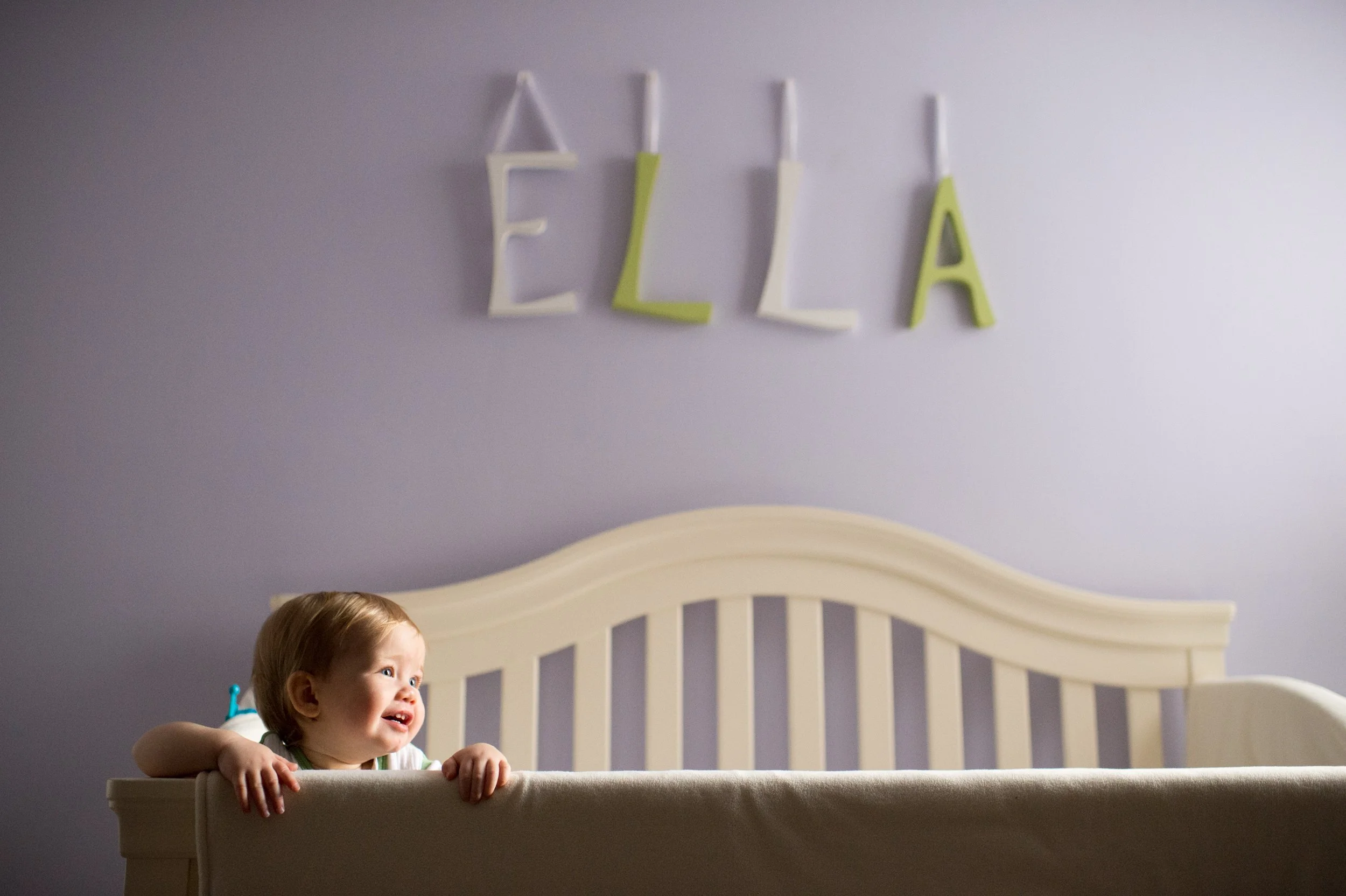 A young girl with light brown hair leaning over a white crib, looking out with a happy expression. Above her, on the wall, there are white and green decorative letters spelling 'ELL'A'.