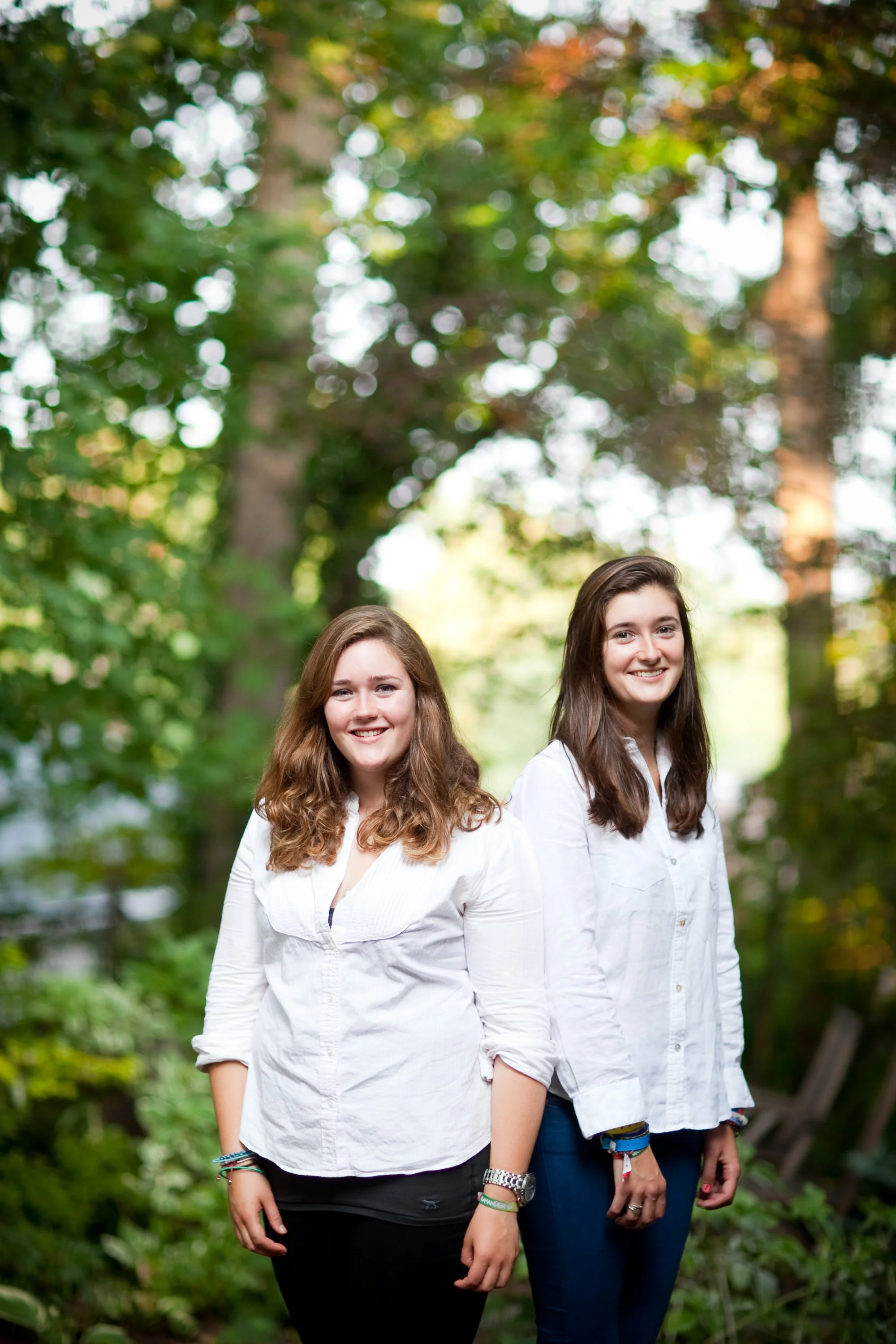 Two young women standing outdoors in a wooded area, smiling at the camera, wearing white shirts and blue jeans.