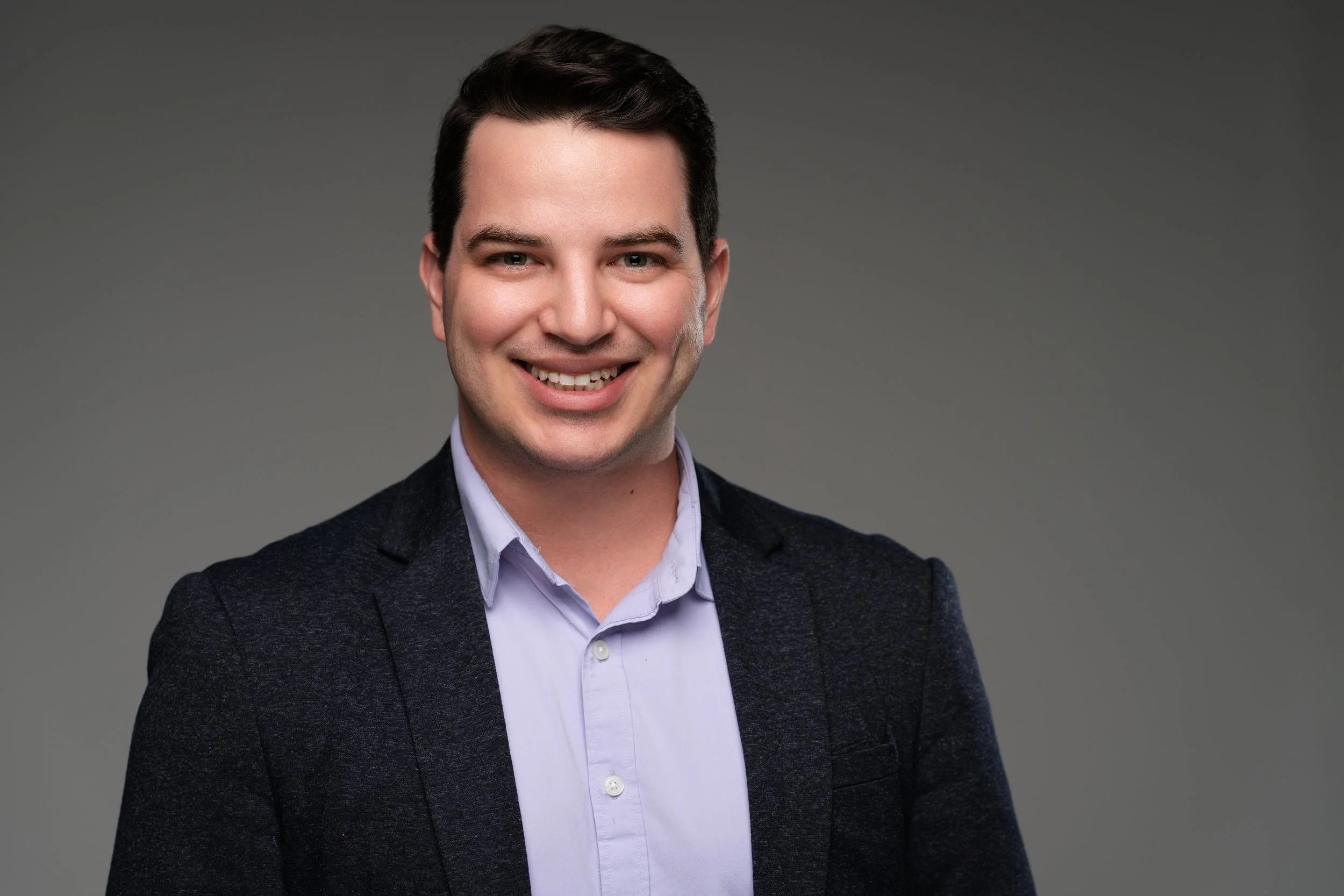 A smiling man with dark hair, wearing a light blue dress shirt and a dark blazer, standing against a plain gray background.