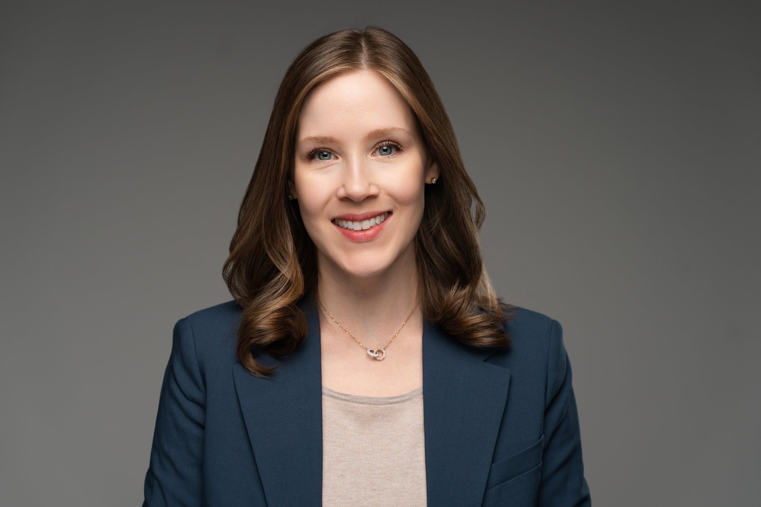 A woman with shoulder-length brown hair, wearing a navy blazer and a beige top, smiling against a gray background.