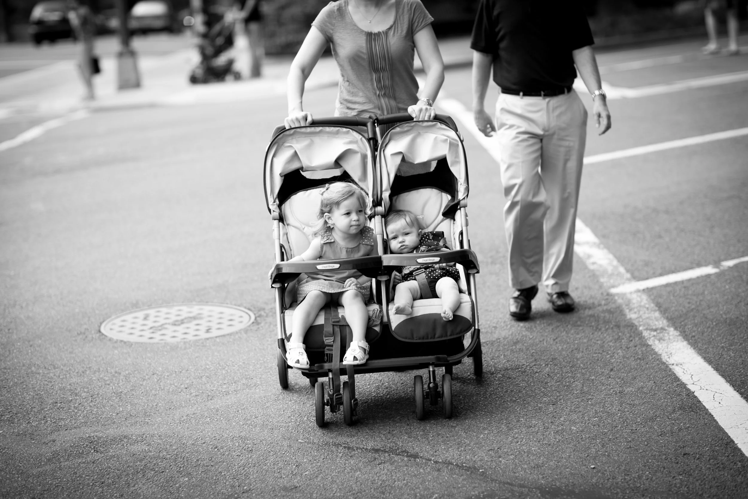 Two young girls sitting in a double stroller being pushed by a woman, with a man walking nearby on a city street.