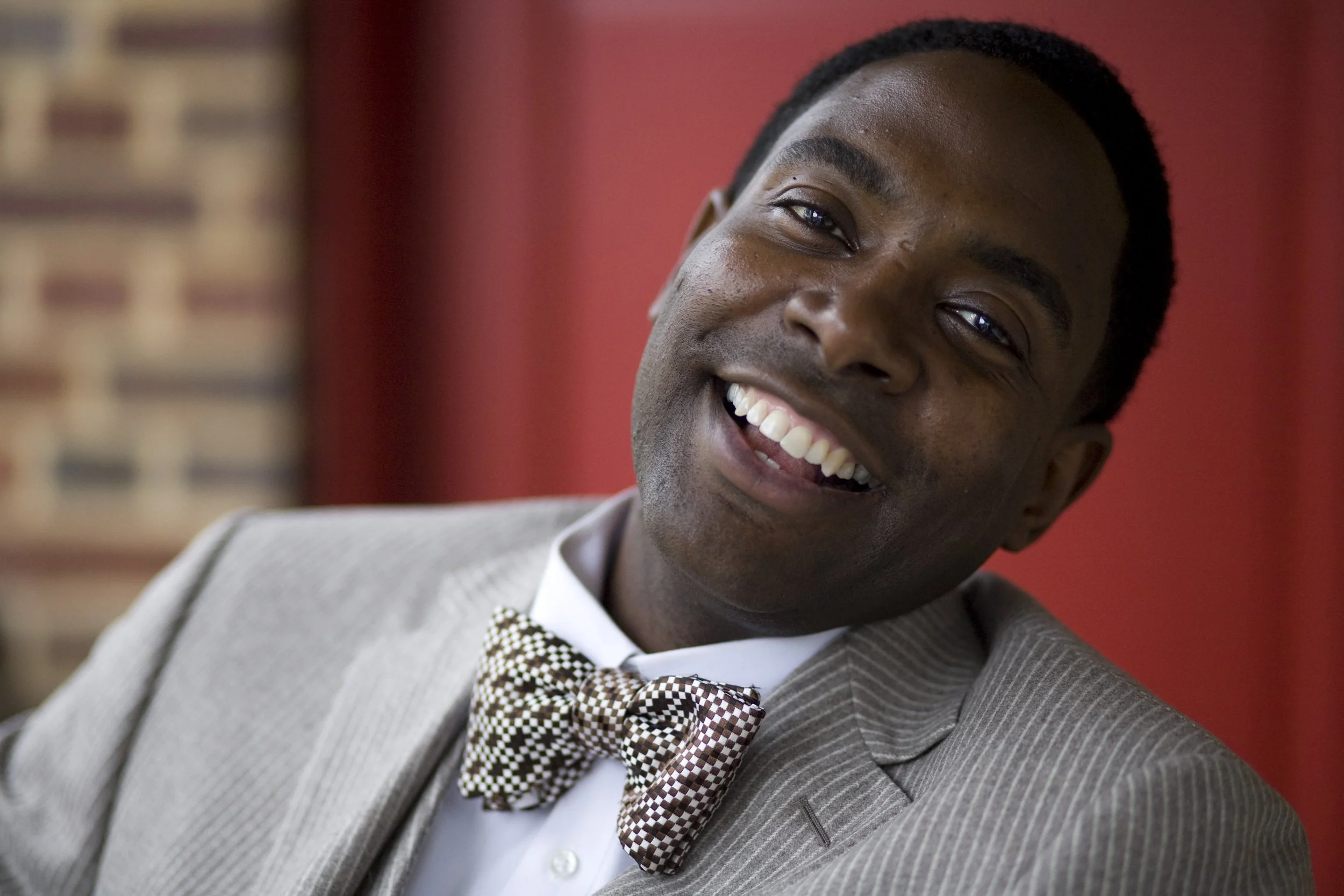 Smiling man in a light gray suit with a patterned bow tie, white shirt, and short hairstyle, indoors with a red curtain in the background.