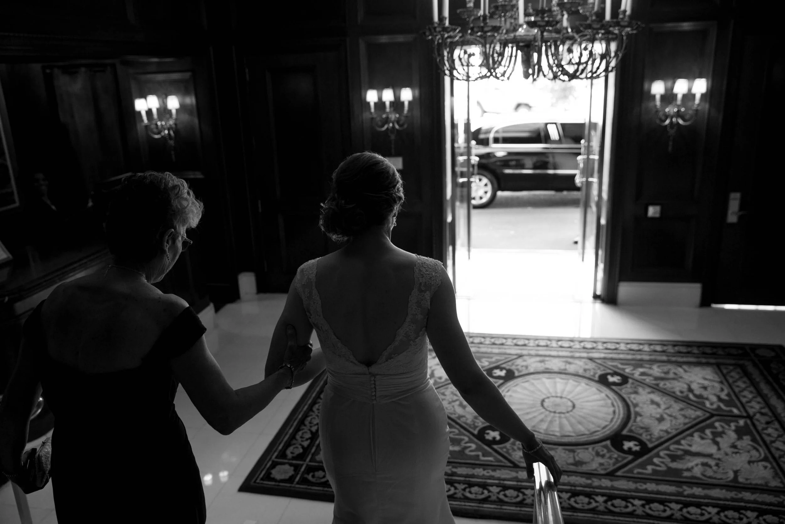 A woman in a wedding dress is descending stairs while being assisted by another woman inside a room with dark wood paneling and a large patterned rug. Light is streaming in from open doors leading outside.
