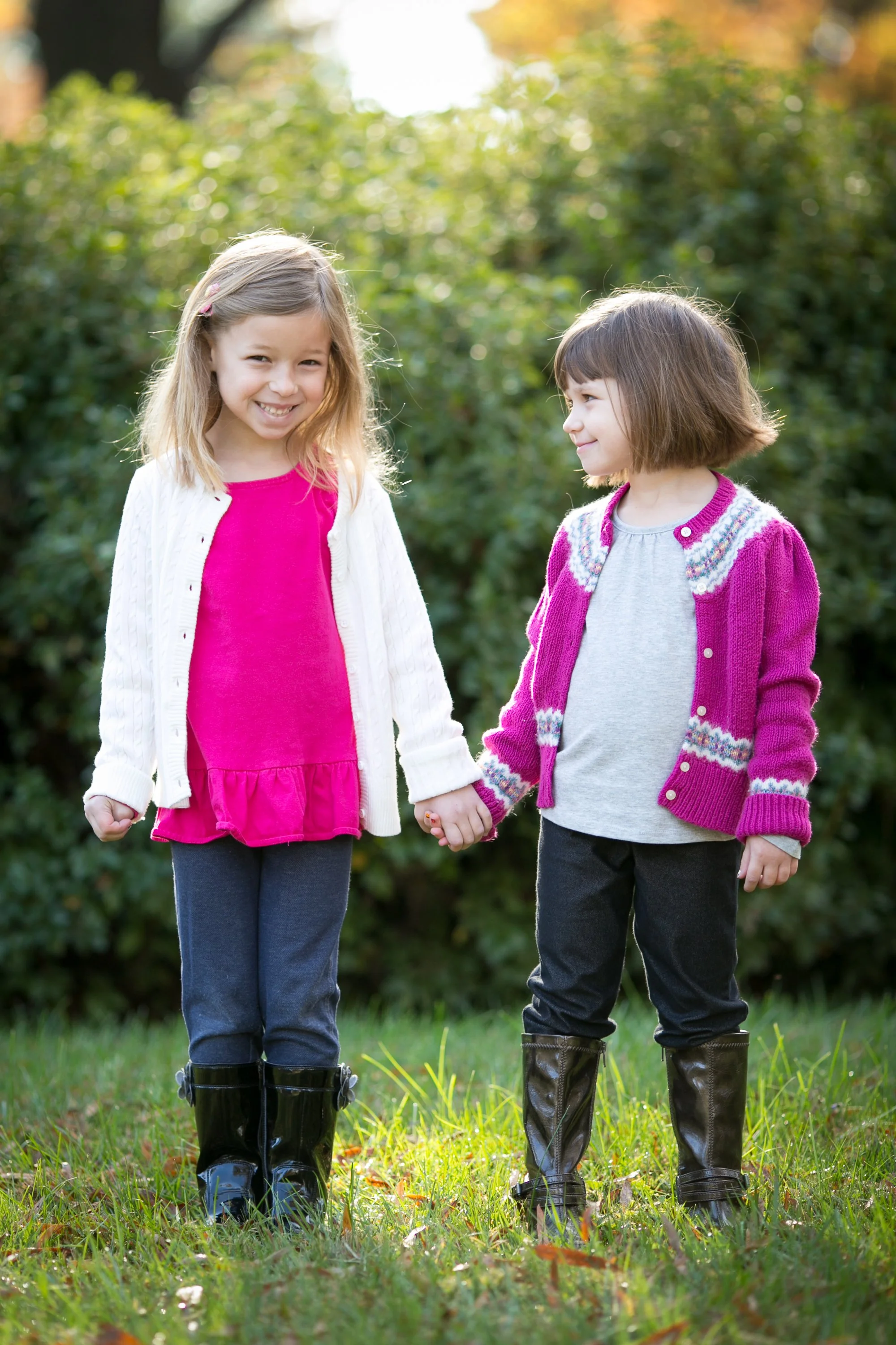 Two smiling young girls holding hands outside in a grassy area with trees in the background, dressed in colorful sweaters and black rain boots.
