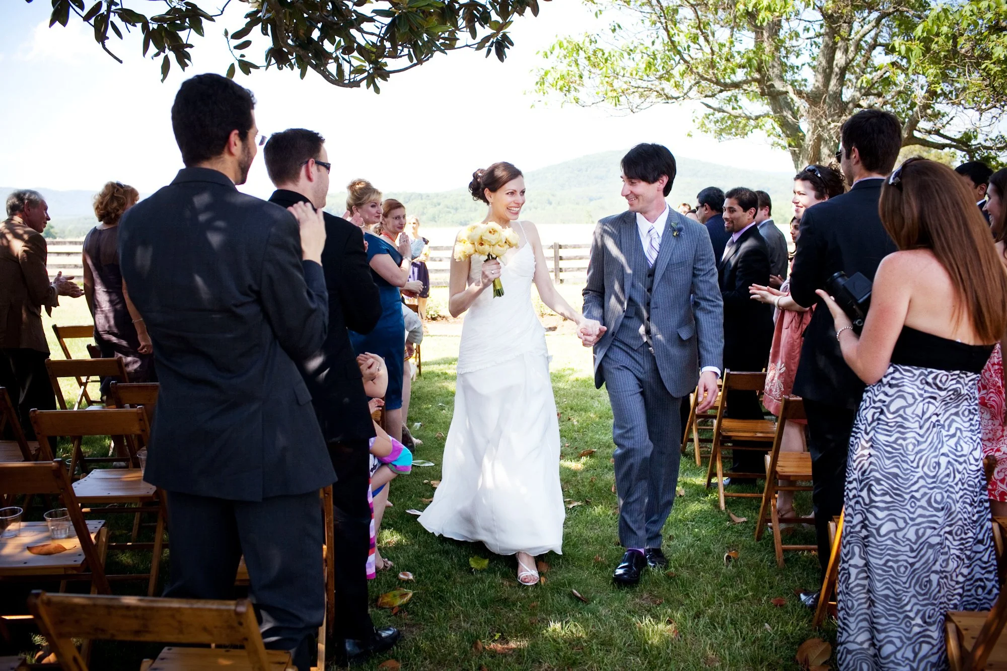A bride and groom walking down the aisle holding hands at their outdoor wedding ceremony, surrounded by guests under a tree with a mountainous landscape in the background.