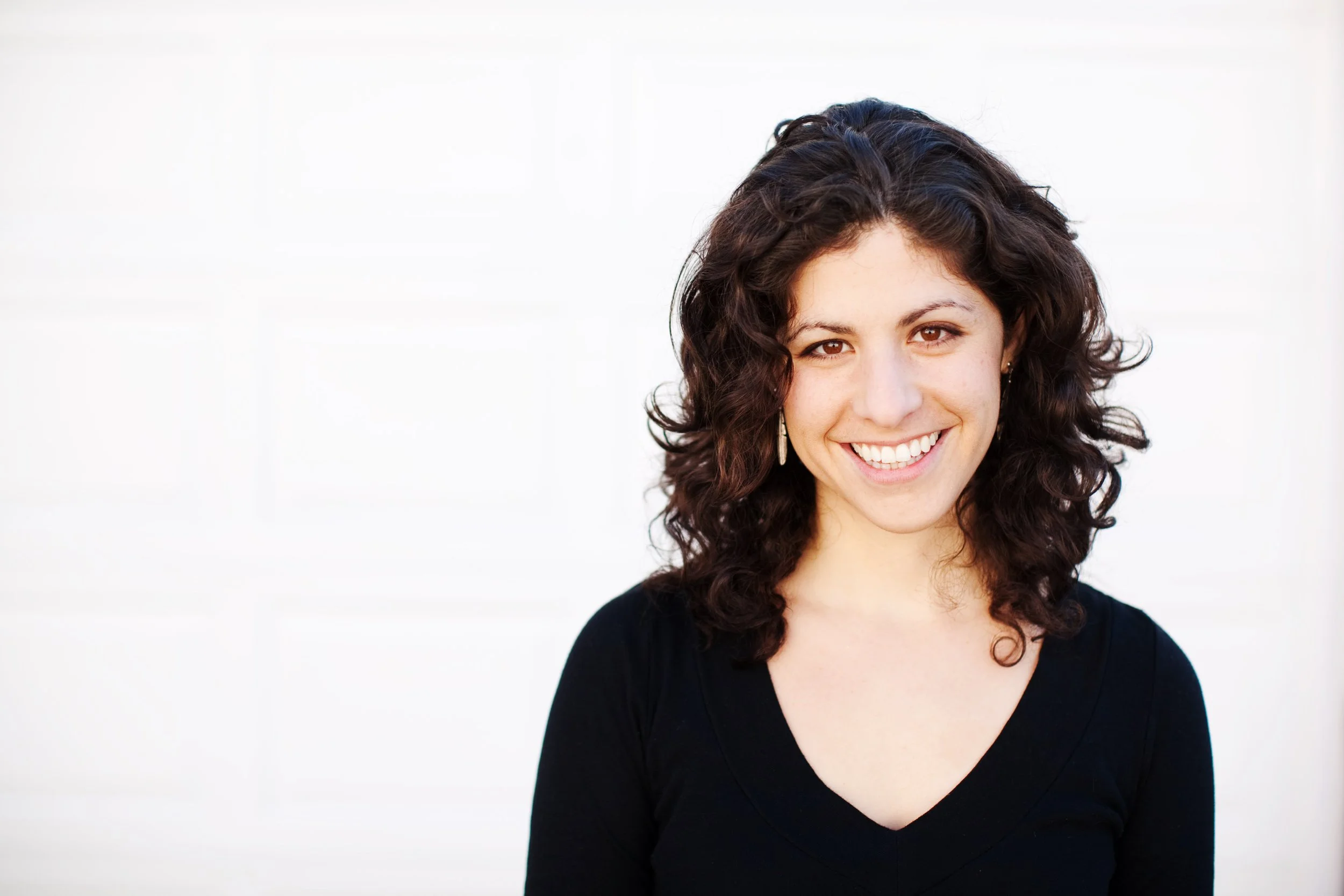 A woman with curly dark brown hair smiling, wearing a black top, standing against a white background.