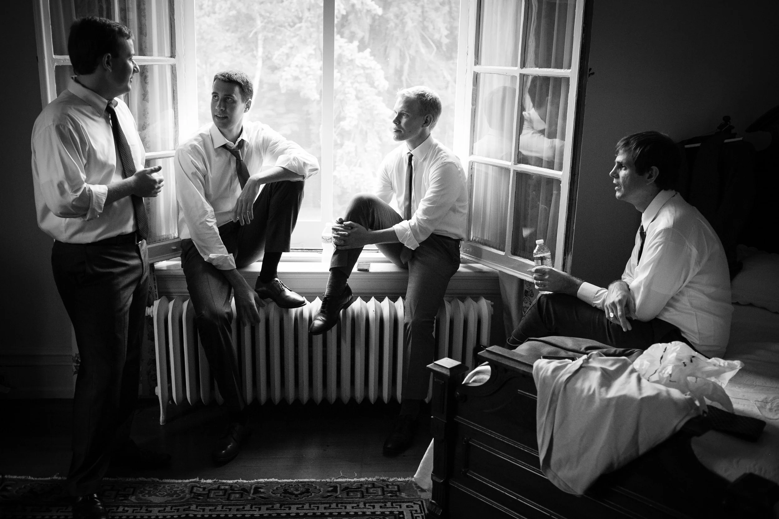 Five men in formal attire wearing dress shirts, ties, and dress shoes are having a conversation in a room with a large window. Four men are sitting on a radiator and windowsill, while one man stands.