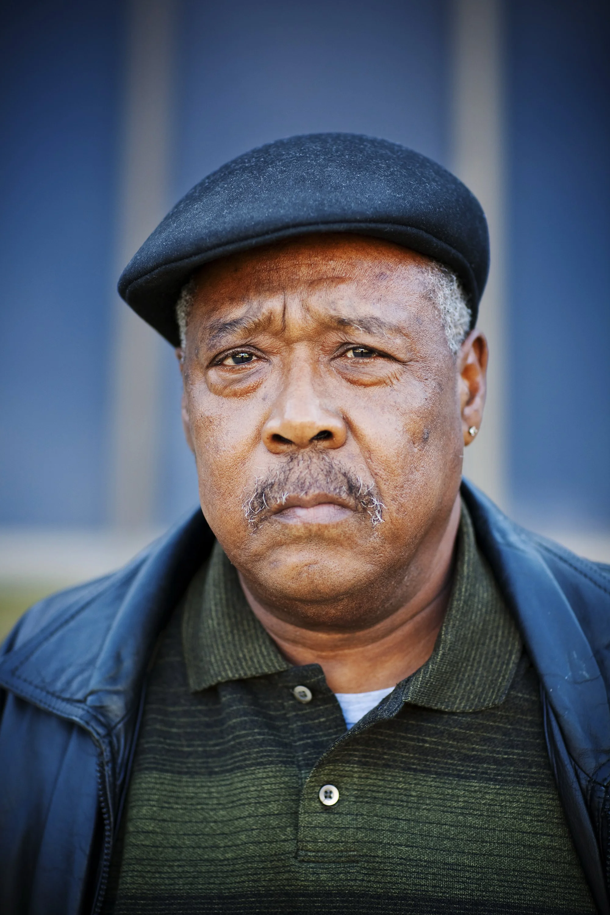 Close-up of an older Black man with gray hair, wearing a black beret, a green collared shirt, and a black jacket, standing outdoors with a blurred background.