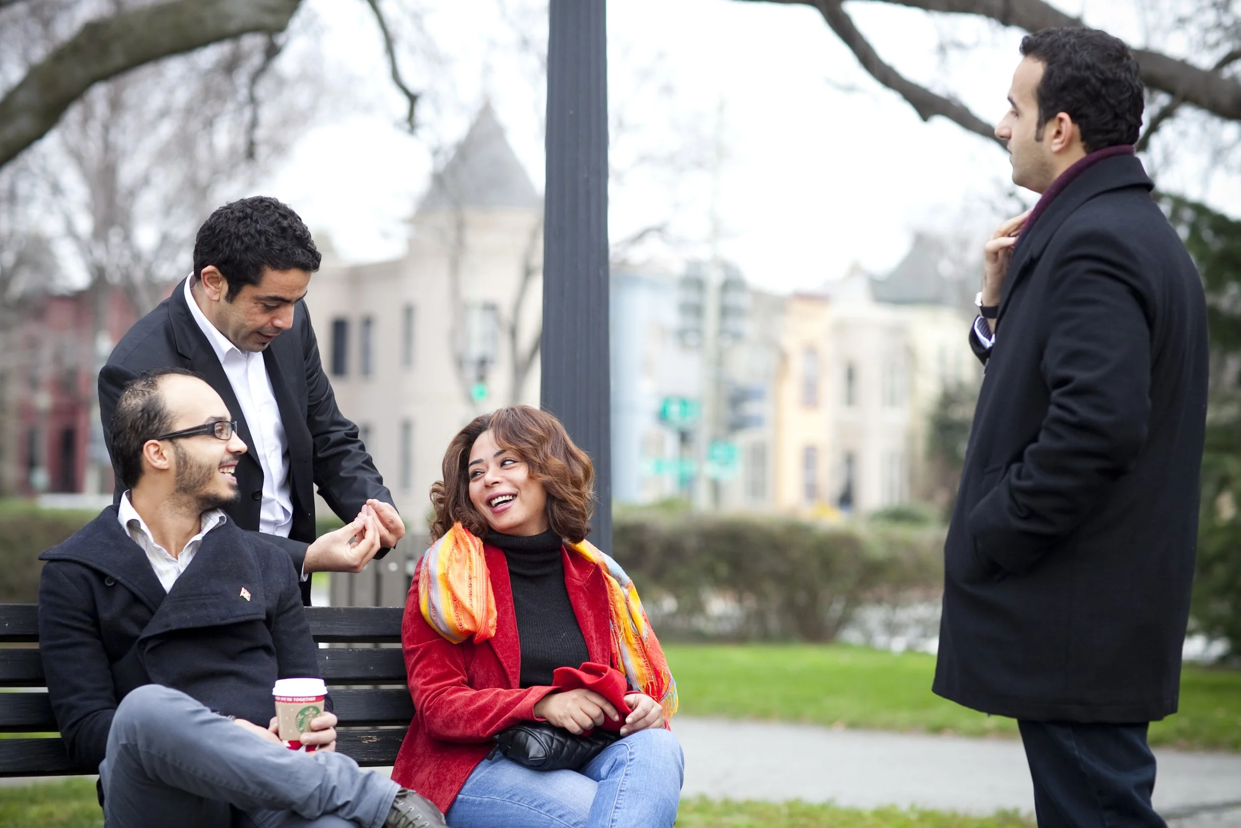 Four friends, two women and two men, are enjoying a conversation at a park. One woman is sitting on a bench with a red coat and colorful scarf, smiling. One man is sitting beside her with a drink, smiling and looking at her. A second man, standing, a