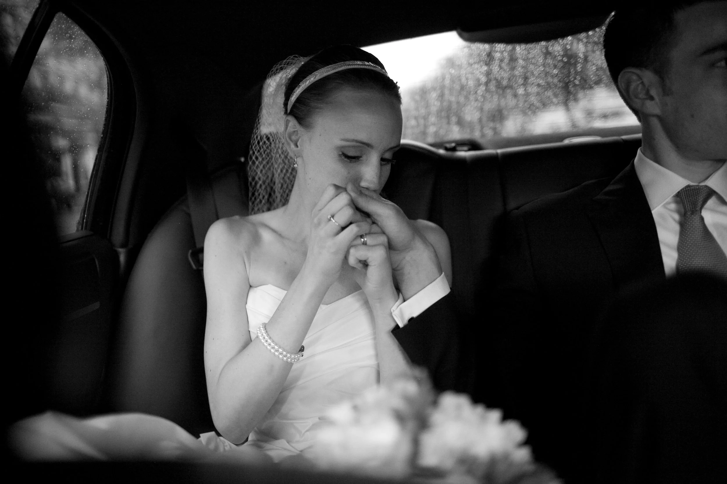 Black and white photo of a bride and groom sitting in the back of a car during a wedding. The bride is holding her hands near her mouth, looking down, with her hair styled in an updo with a veil. The groom is sitting next to her, dressed in a suit an