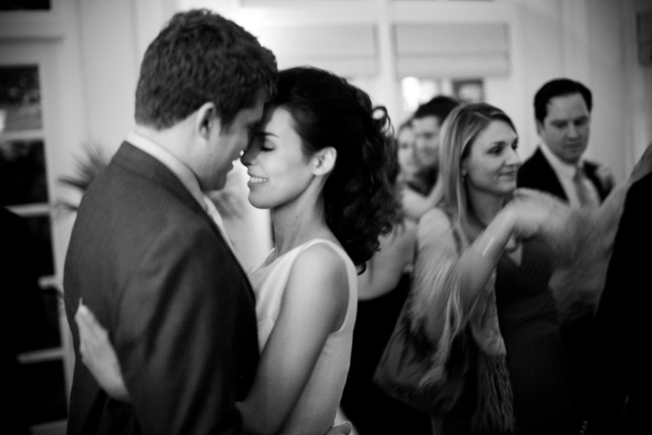 A black and white photo of a smiling couple dancing closely with their foreheads touching, surrounded by other people at a social event.
