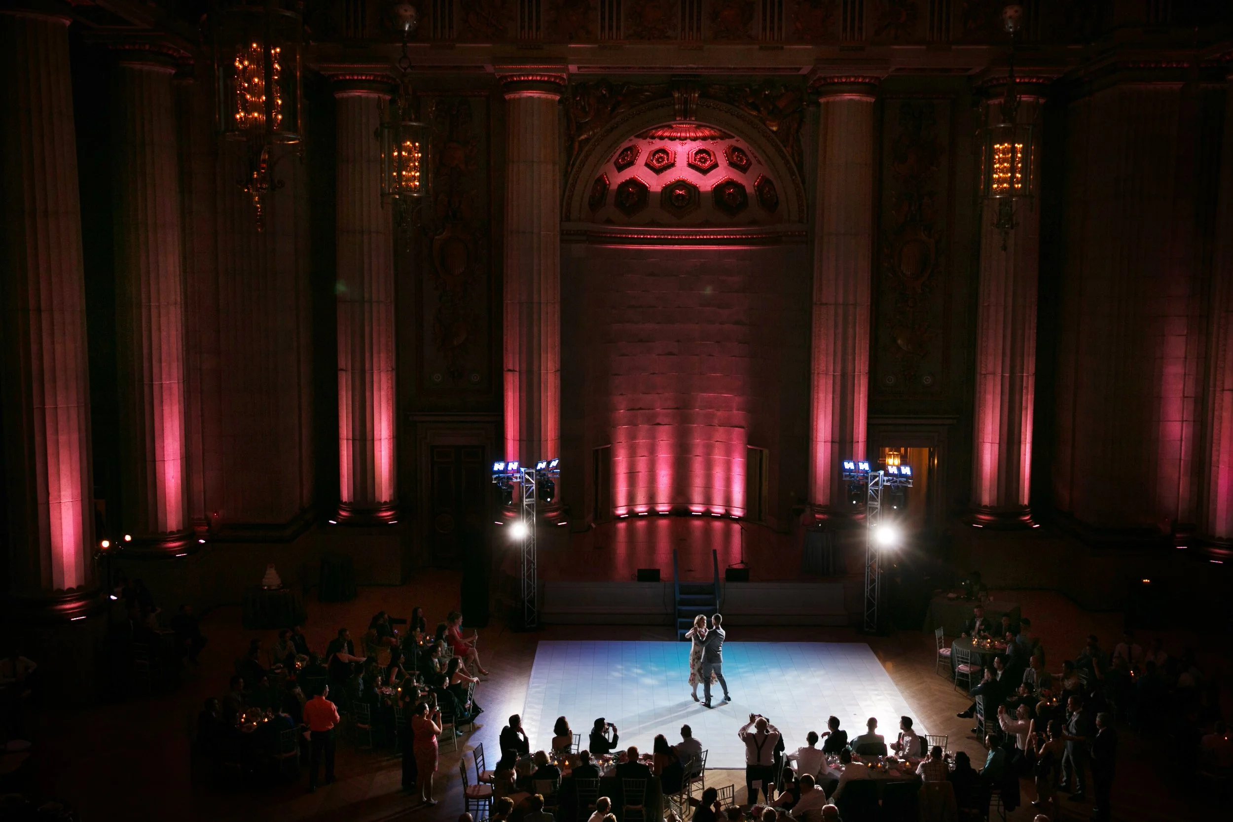 A couple dancing in the center of a grand ballroom with pink lighting, surrounded by seated guests at tables.