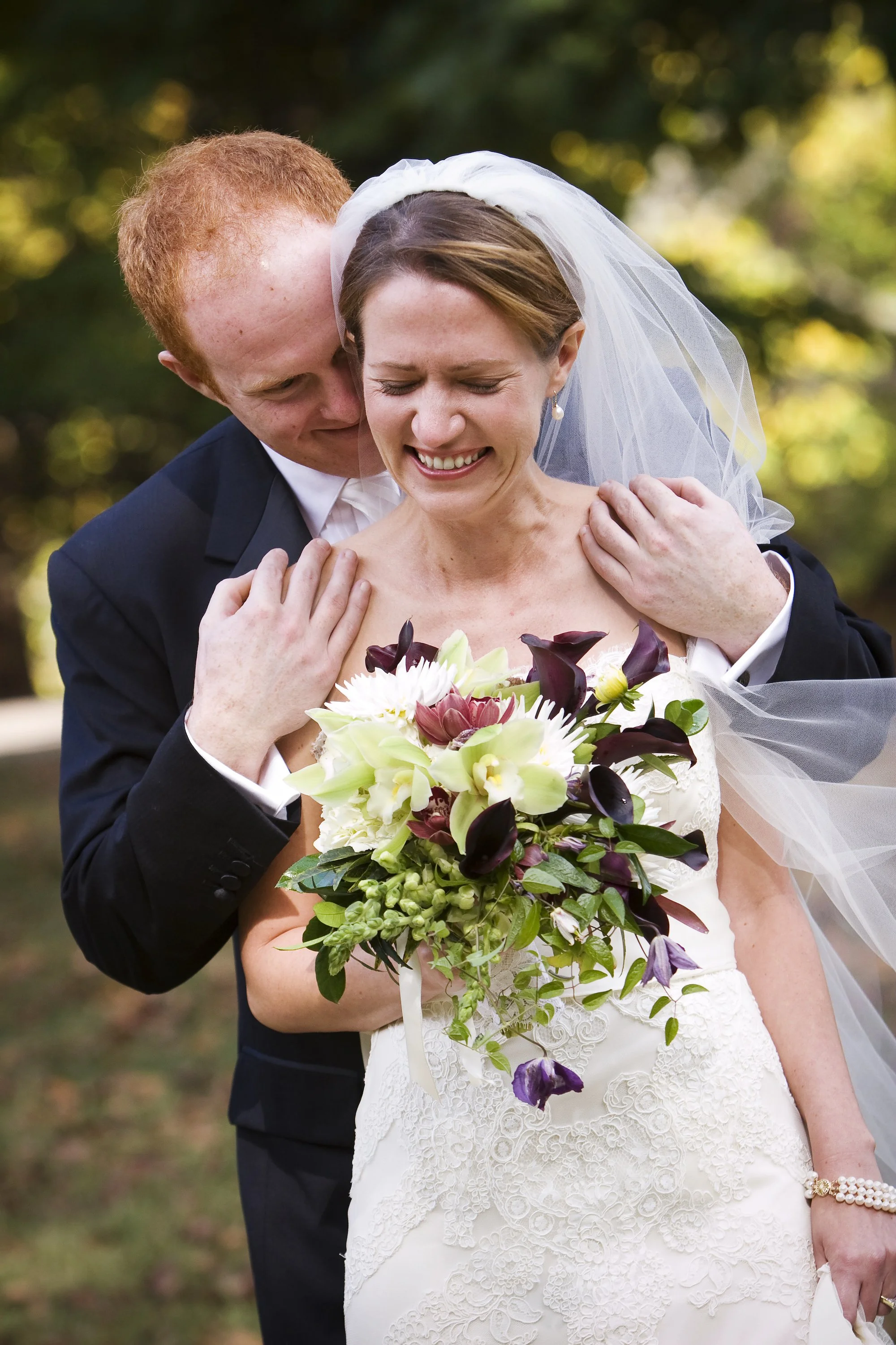 A bride and groom in wedding attire sharing an affectionate moment outdoors, with the groom embracing the bride from behind while she holds a bouquet of flowers.