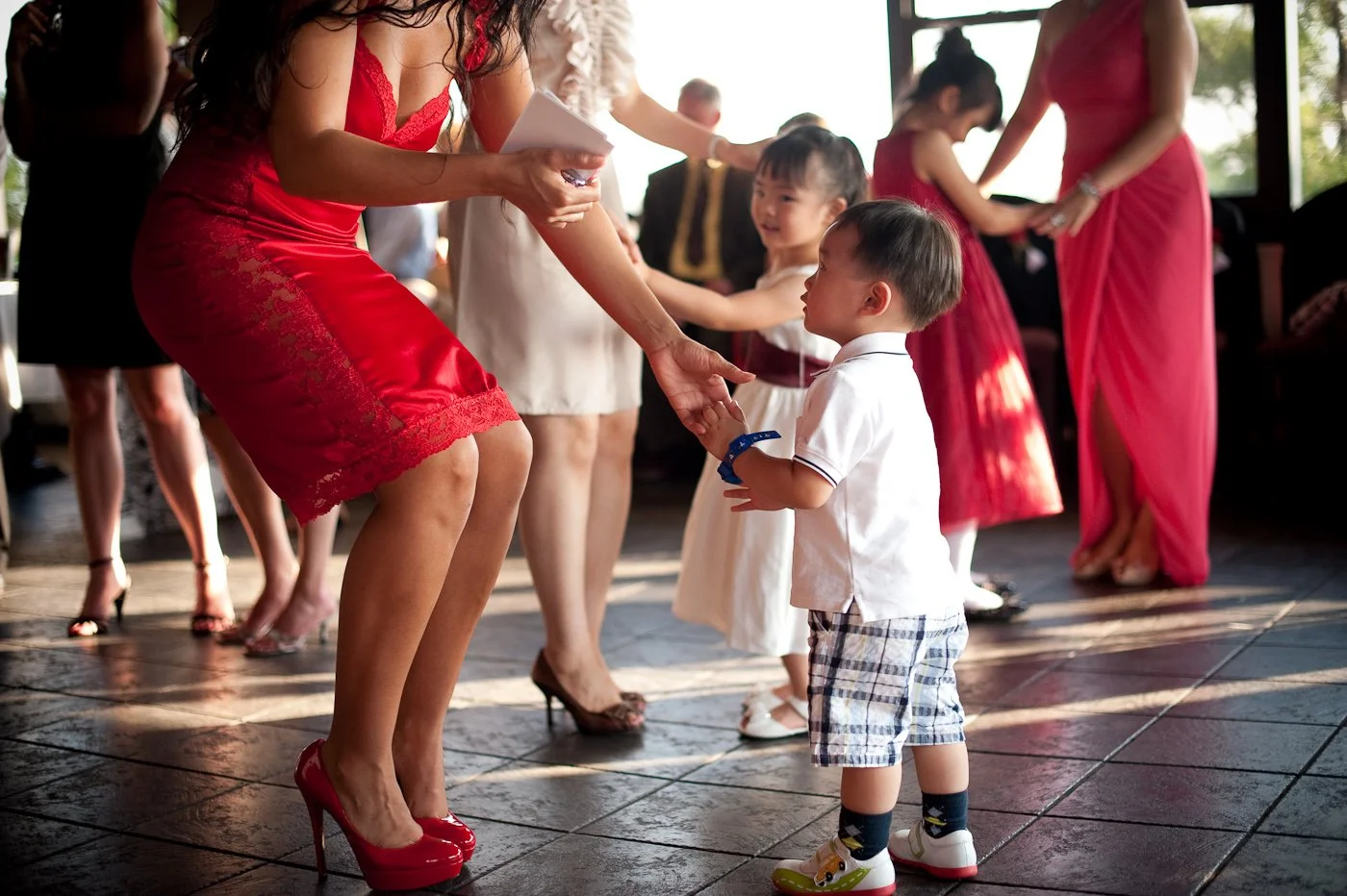 A woman in a red dress and high heels is handing a certificate or award to a young boy in a white shirt and plaid shorts at a celebration or party, with other children and women in red dresses dancing or interacting in the background.