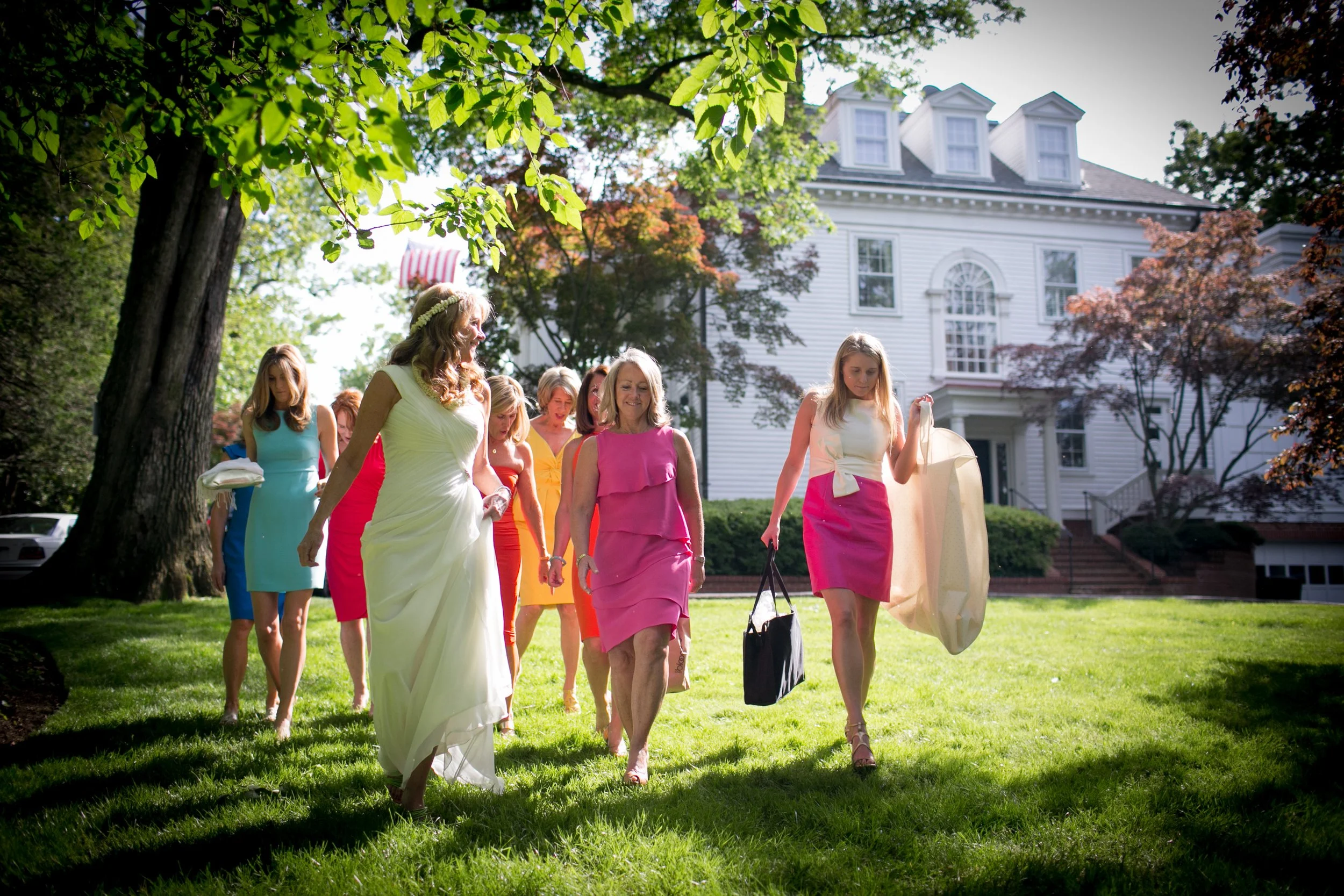 Group of women in colorful dresses walking on a lush green lawn in front of a large white house with trees around on a sunny day.