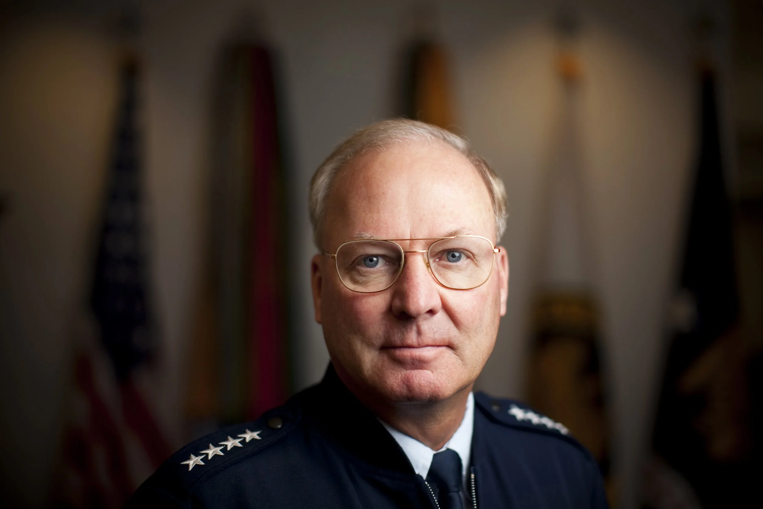 A four star general in military uniform, with glasses and short blond hair, stands in front of a blurred background displaying flags.