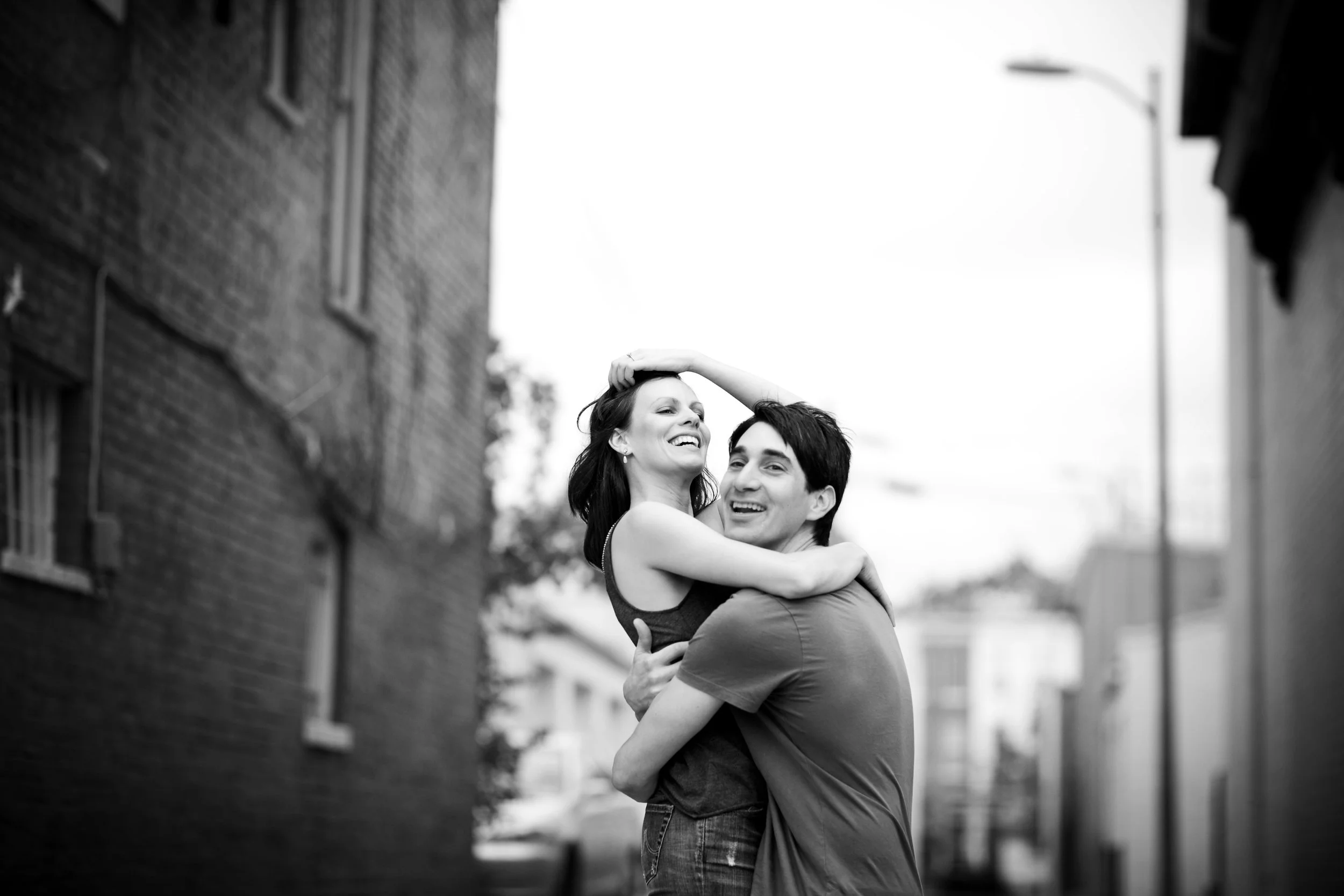 A joyful couple embracing and smiling in an outdoor alleyway with brick buildings, black and white photo.