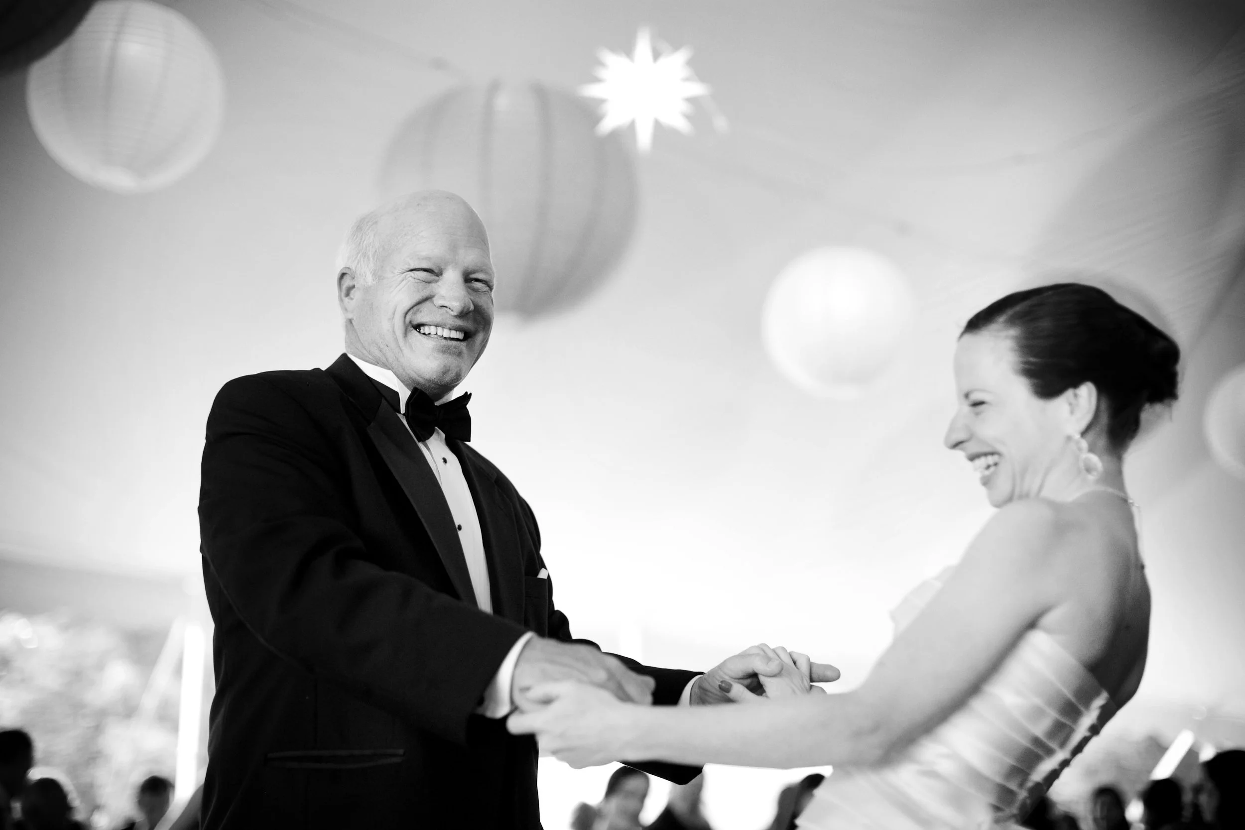 A couple dancing at their wedding reception, with the man wearing a tuxedo and the woman in a strapless dress, smiling and holding hands, decorated with hanging lanterns and a star-shaped ornament.