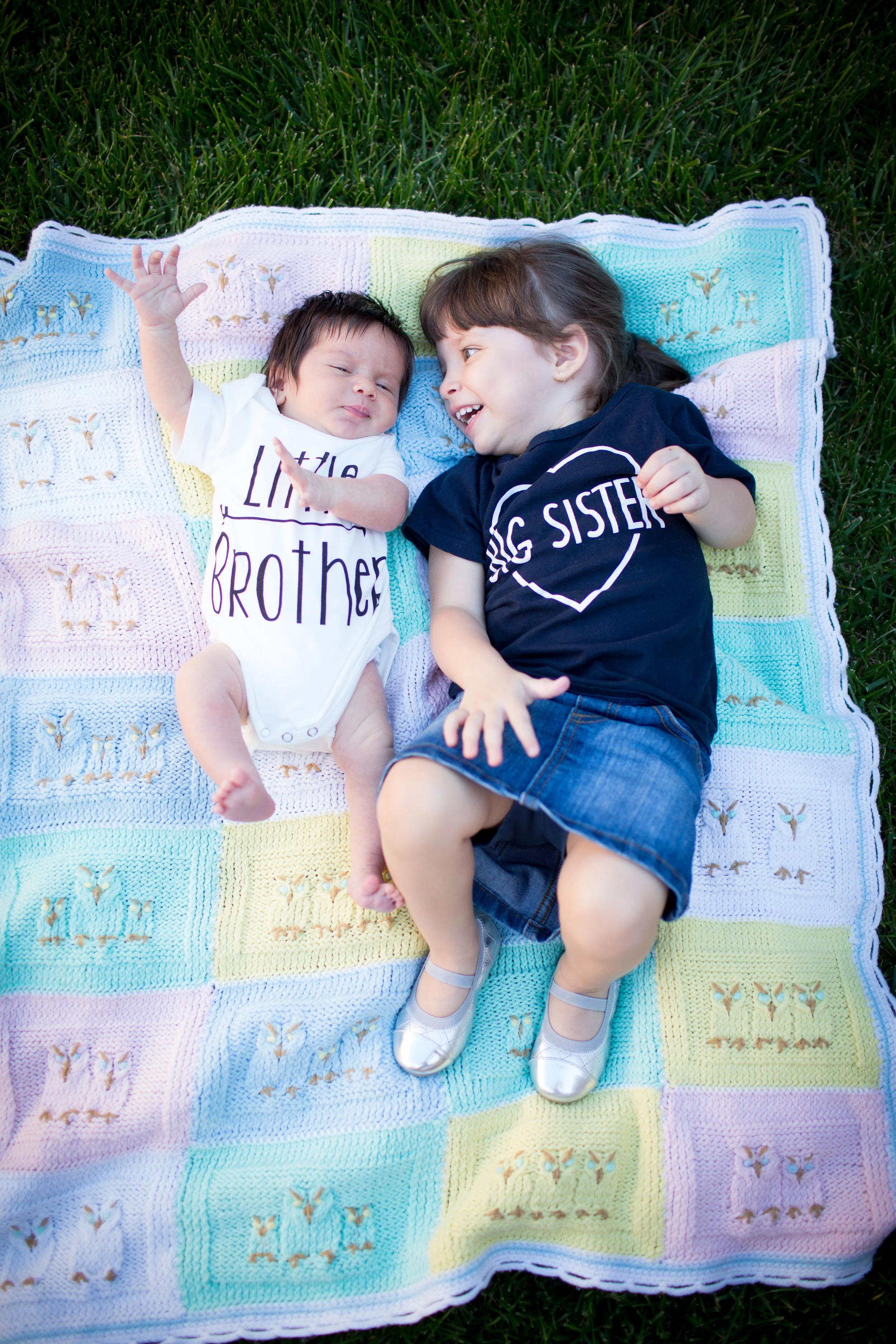 Two young girls, one in a white shirt and one in a navy t-shirt, lying on a colorful crocheted blanket outdoors on grass, smiling and playing together.