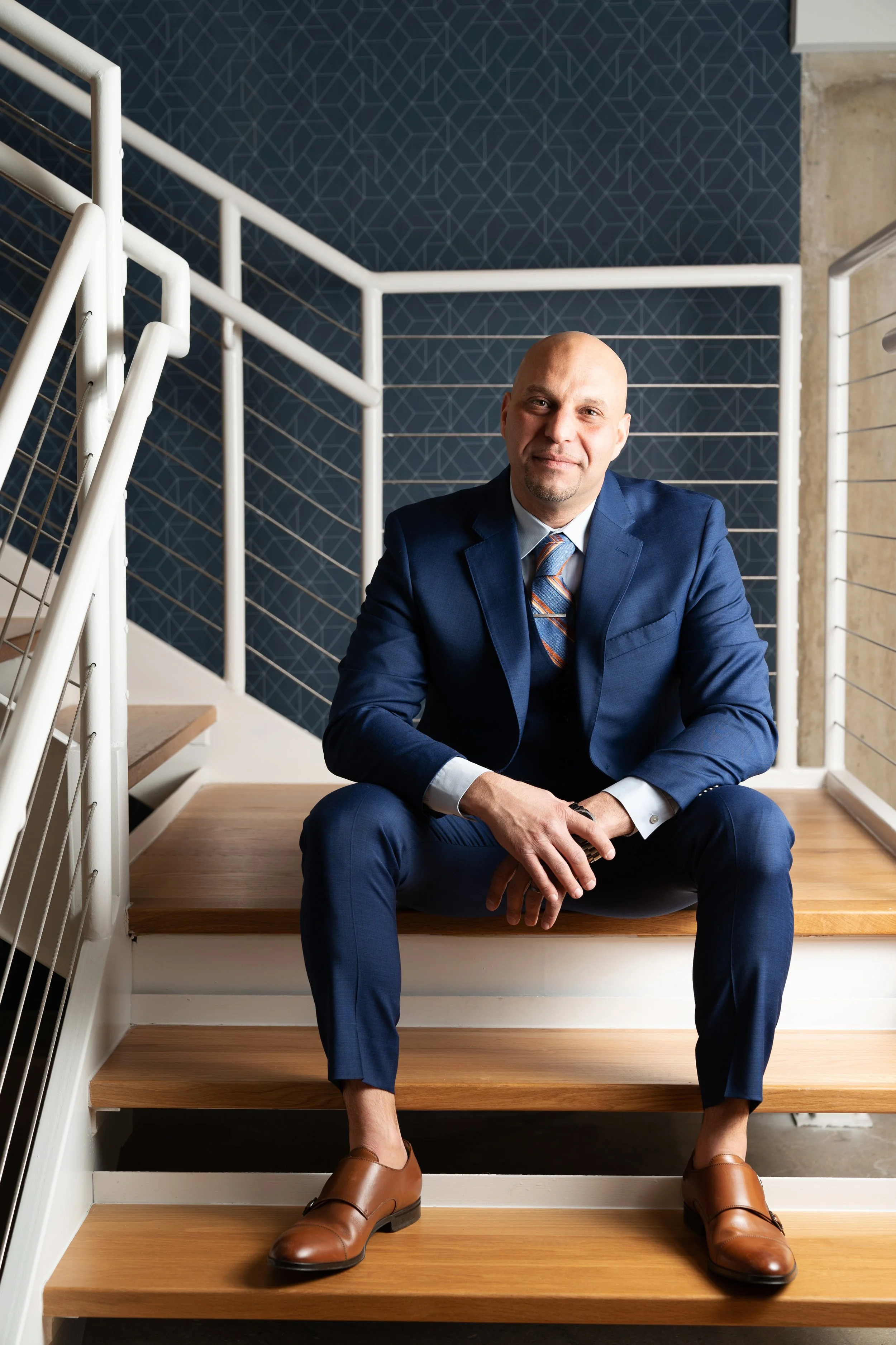A man in a blue suit sitting on wooden stairs, smiling, with a modern railing and geometric-patterned wall in the background.