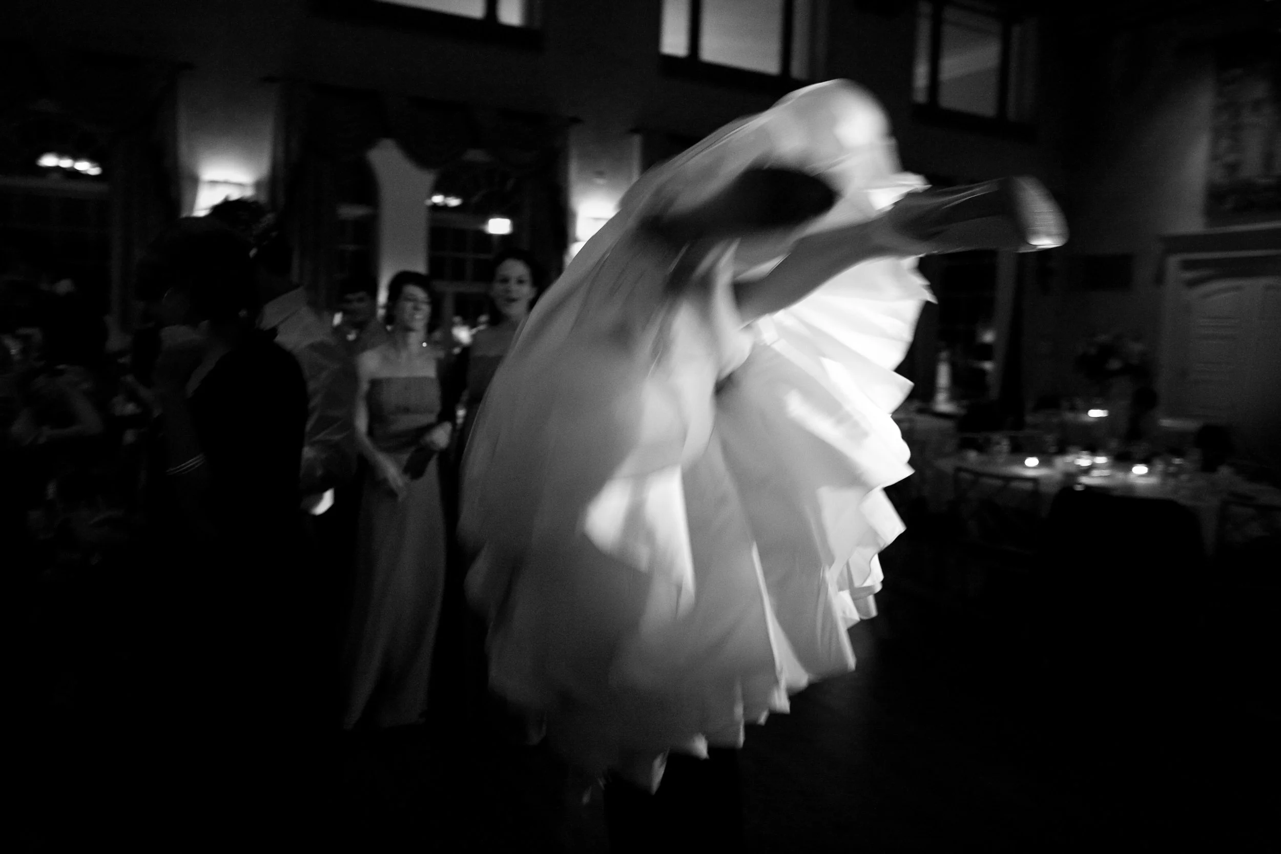 Black and white photo of a bride twirling in her wedding dress at a wedding reception, with guests watching in the background inside a decorated venue.