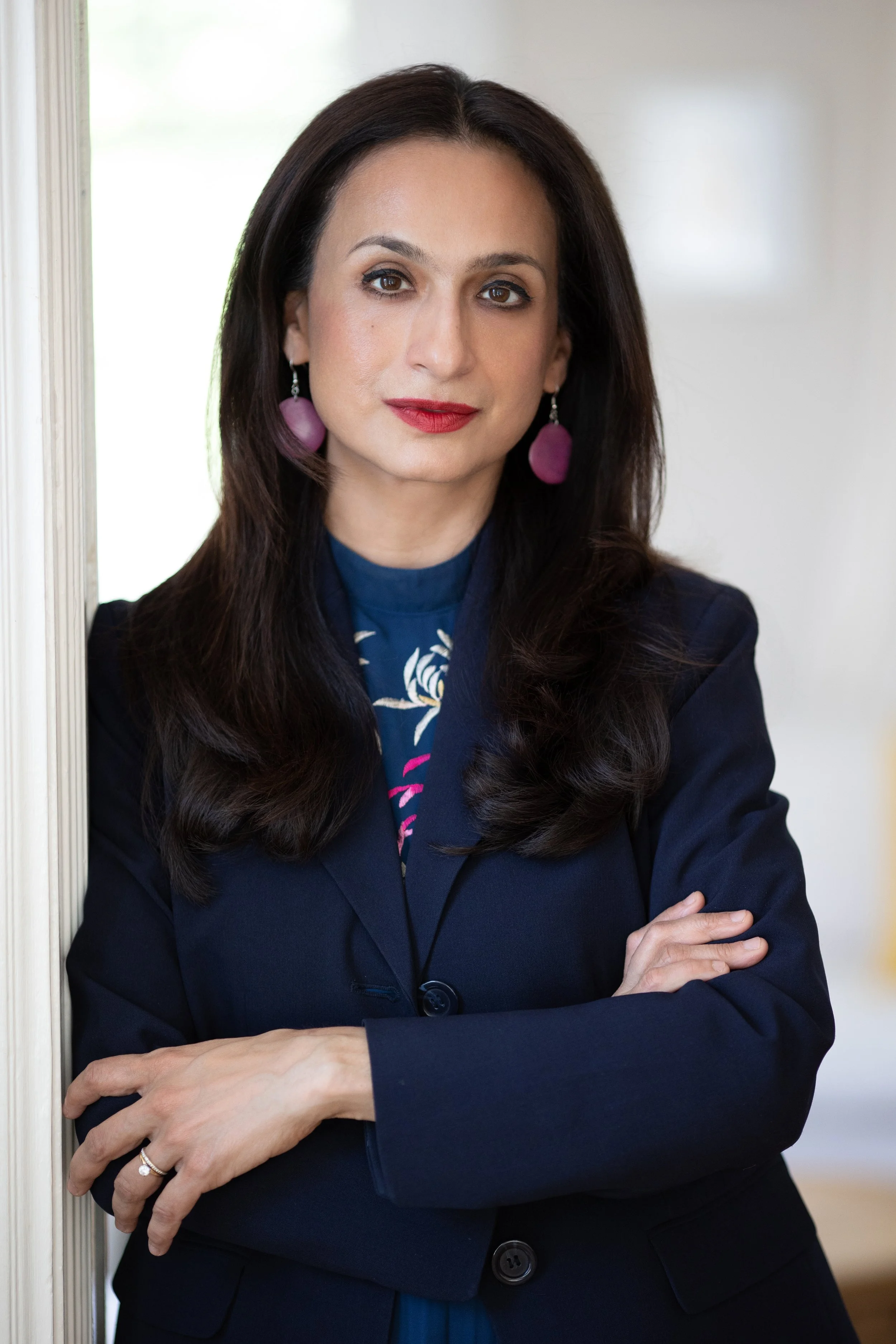 A woman with long dark hair, wearing a dark blazer and purple earrings, standing with her arms crossed and leaning against a wall, looking at the camera.
