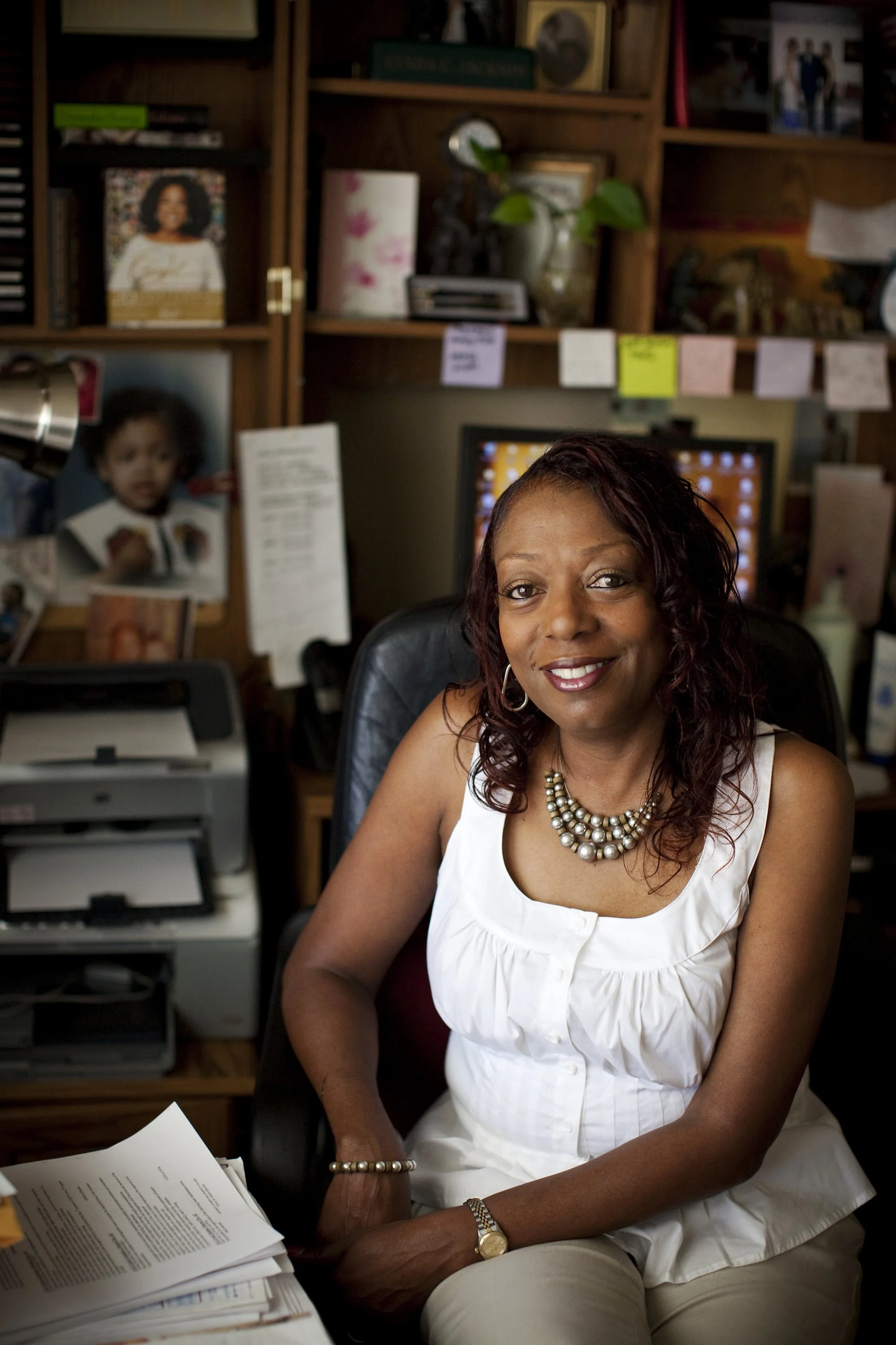 A woman with auburn hair wearing a white sleeveless top and pearl jewelry sitting at a cluttered office desk.