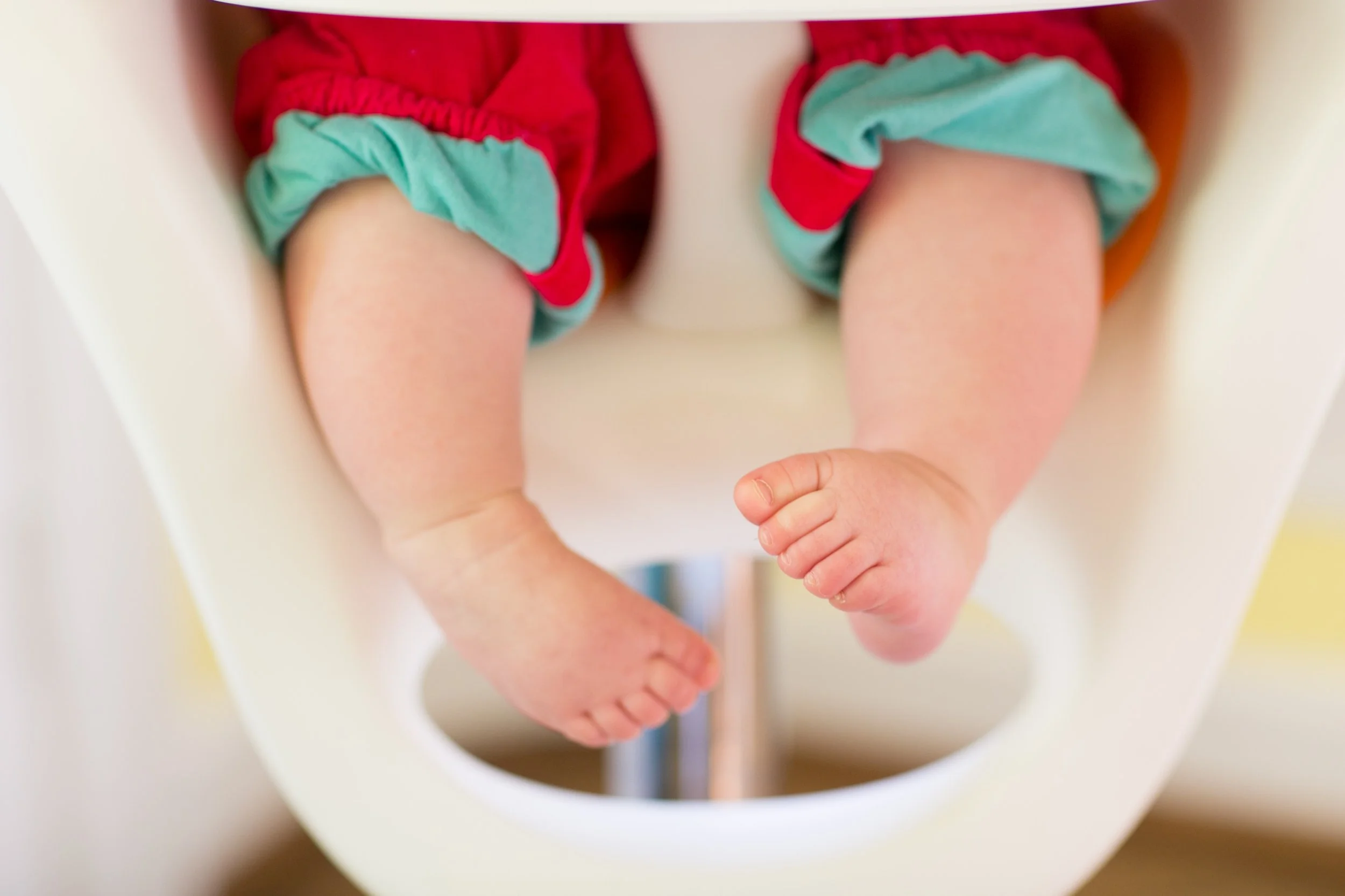 Close-up of a baby's legs and feet as they sit in a white highchair, wearing red and teal clothing.