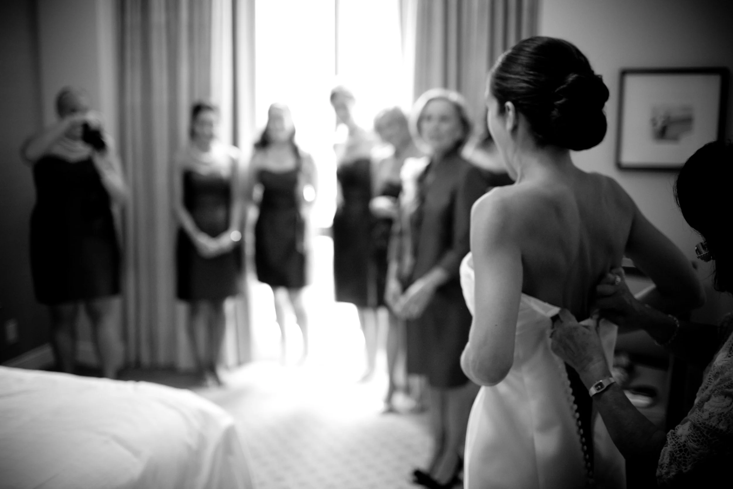 A bride in a strapless wedding dress gets ready, with her back to the camera, while women gather in a hotel room.